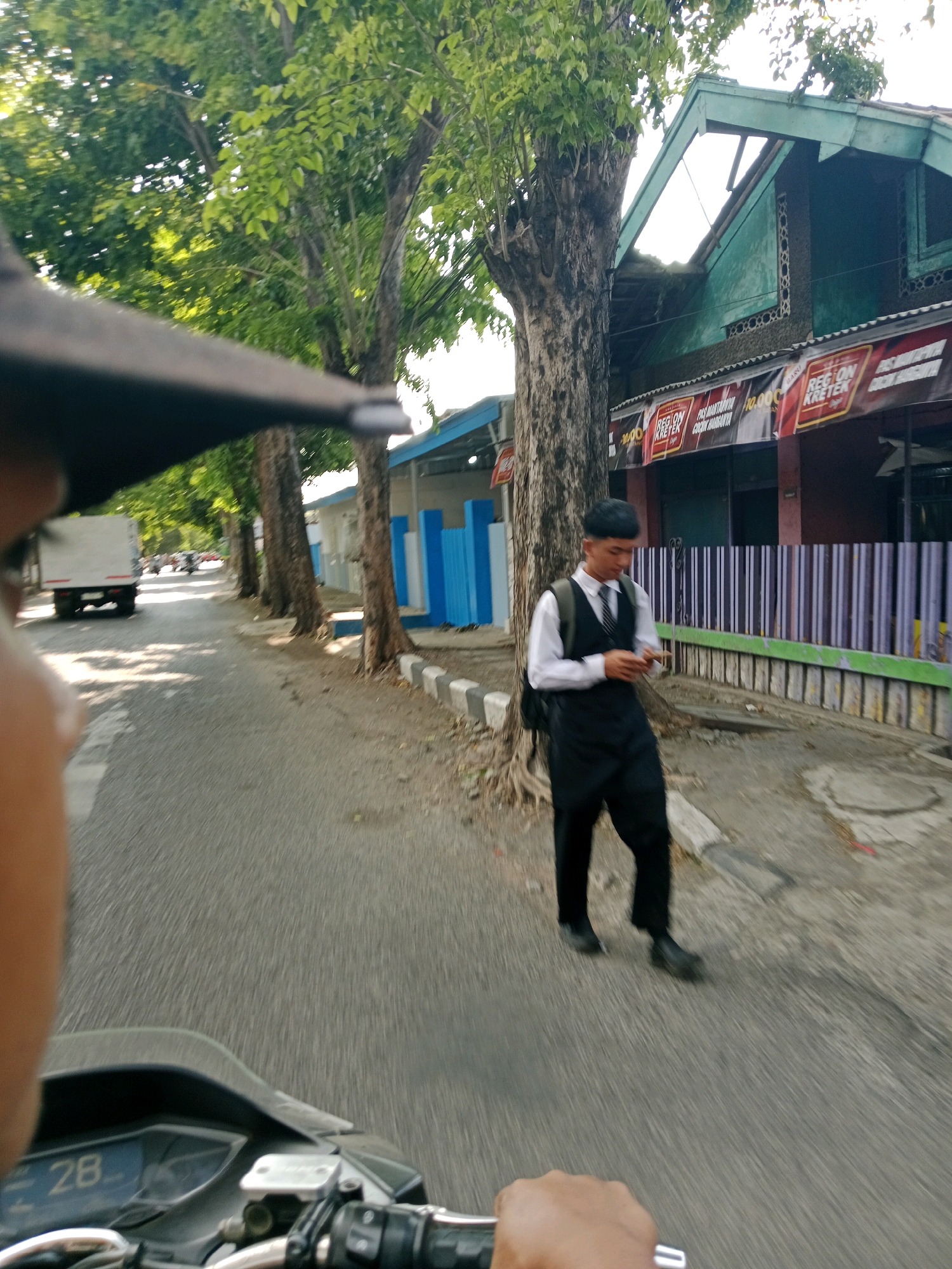In Probolinggo, Indonesia, a young man in a formal outfit comprising a white shirt, black vest, and black pants, possibly a waiter or a student, walks along a roadside. He is holding a mobile phone and appears to be looking at it. The scene unfolds next to a row of trees lining a paved road. In the background, a building with a distinctive green-tiled roof and a striped fence is visible, along with a white truck and motorcycles on the road, suggesting a moderately busy urban environment. The image is captured from the perspective of someone on a motorcycle, as indicated by the handlebars and speedometer showing "28" in the foreground.