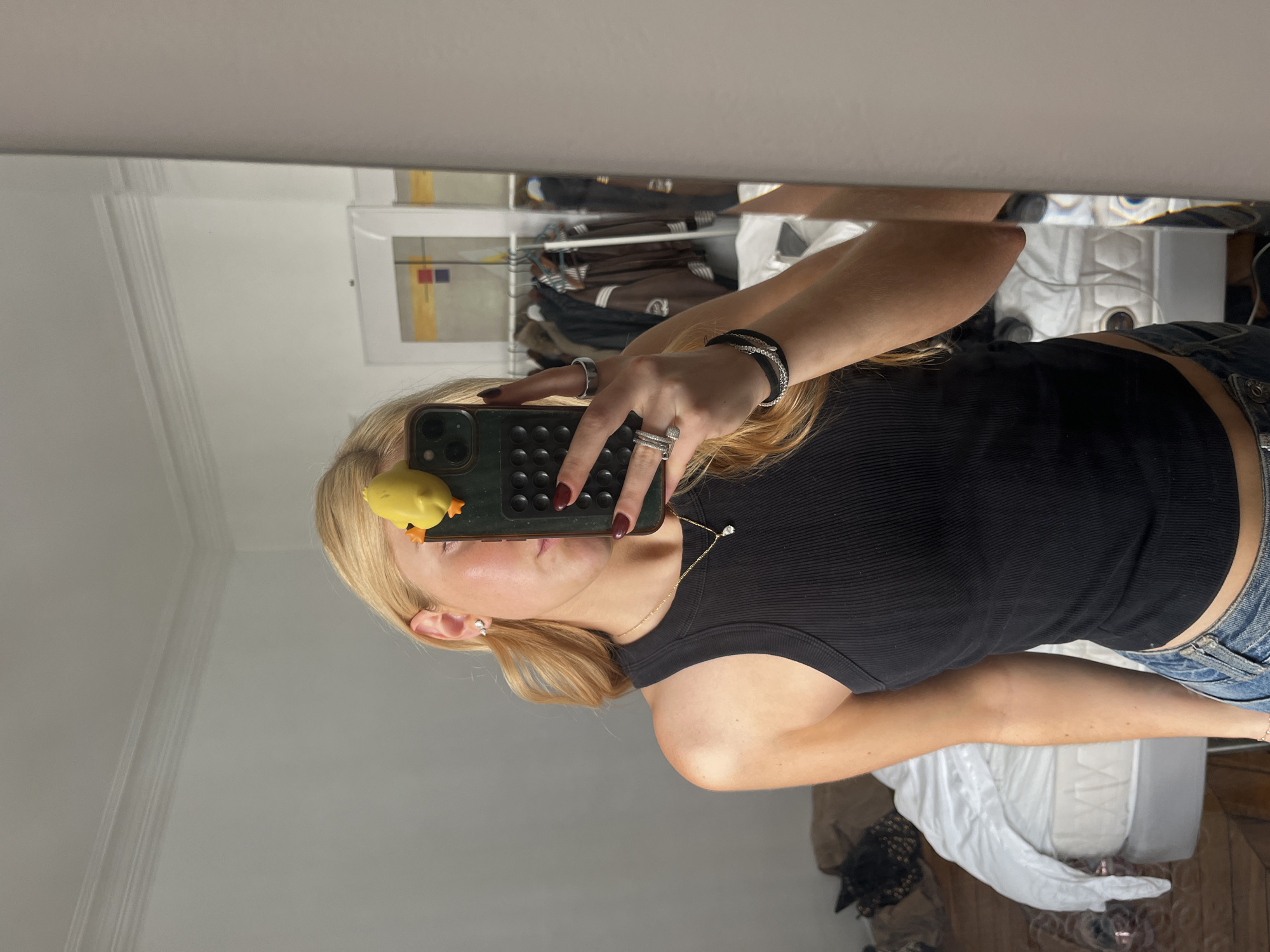 The image is a selfie taken in front of a mirror. A person with blond hair is holding a phone, with a small yellow rubber duck attached to the phone. The person is wearing a black tank top and blue jeans. The background shows a room interior, including a closet with clothes hanging inside, a painting, a bed with white sheets, and a wooden floor. There is no indication that this image was taken in Paris, France.