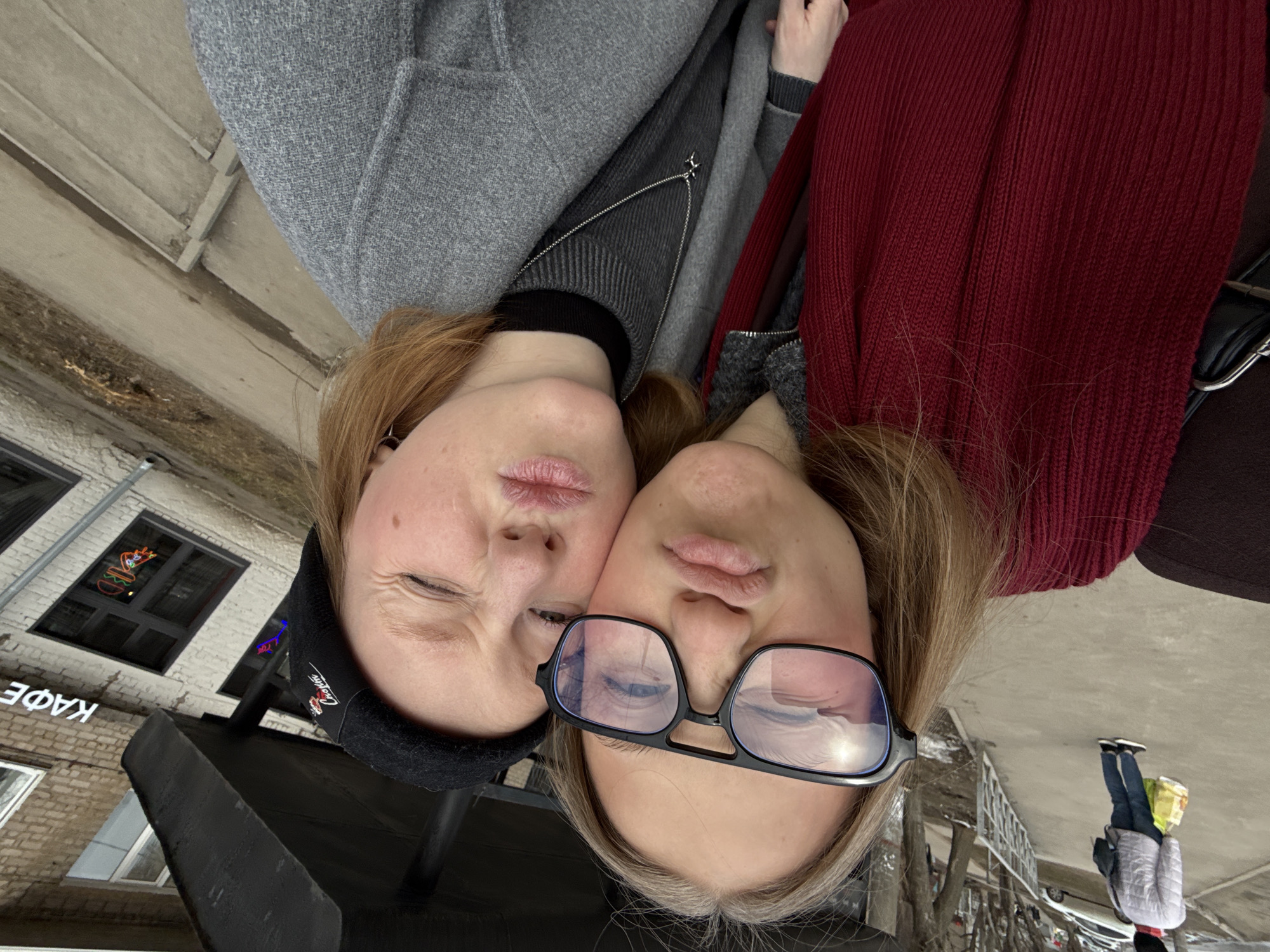 Two young women pose for a selfie. The woman on the left wears a black beanie and a gray coat. The woman on the right wears glasses and a maroon cardigan. They stand outside a building with Cyrillic lettering "КАФЕ" indicating "CAFE". Neon signs adorn the windows. The location appears to be in Perm, Russia. A passerby is visible in the background.