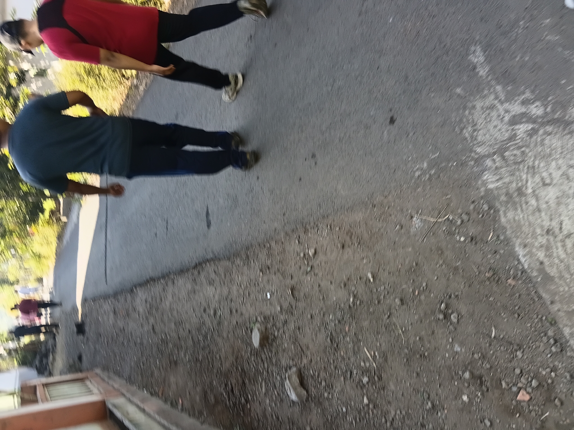 In Tanggul Wetan, Indonesia, two individuals are walking away from the camera on a paved road. The person in the foreground on the right wears a red shirt and black pants, with a black cap. The person to their left wears a dark blue t-shirt and dark blue pants. Behind them, several other figures are visible in the distance, also walking on the road. The environment includes a dirt shoulder on the left side of the road with some scattered rocks and debris, and vegetation and trees lining the right side of the road. A building with an orange facade is partially visible on the far left of the image. The lighting suggests it is daytime.
