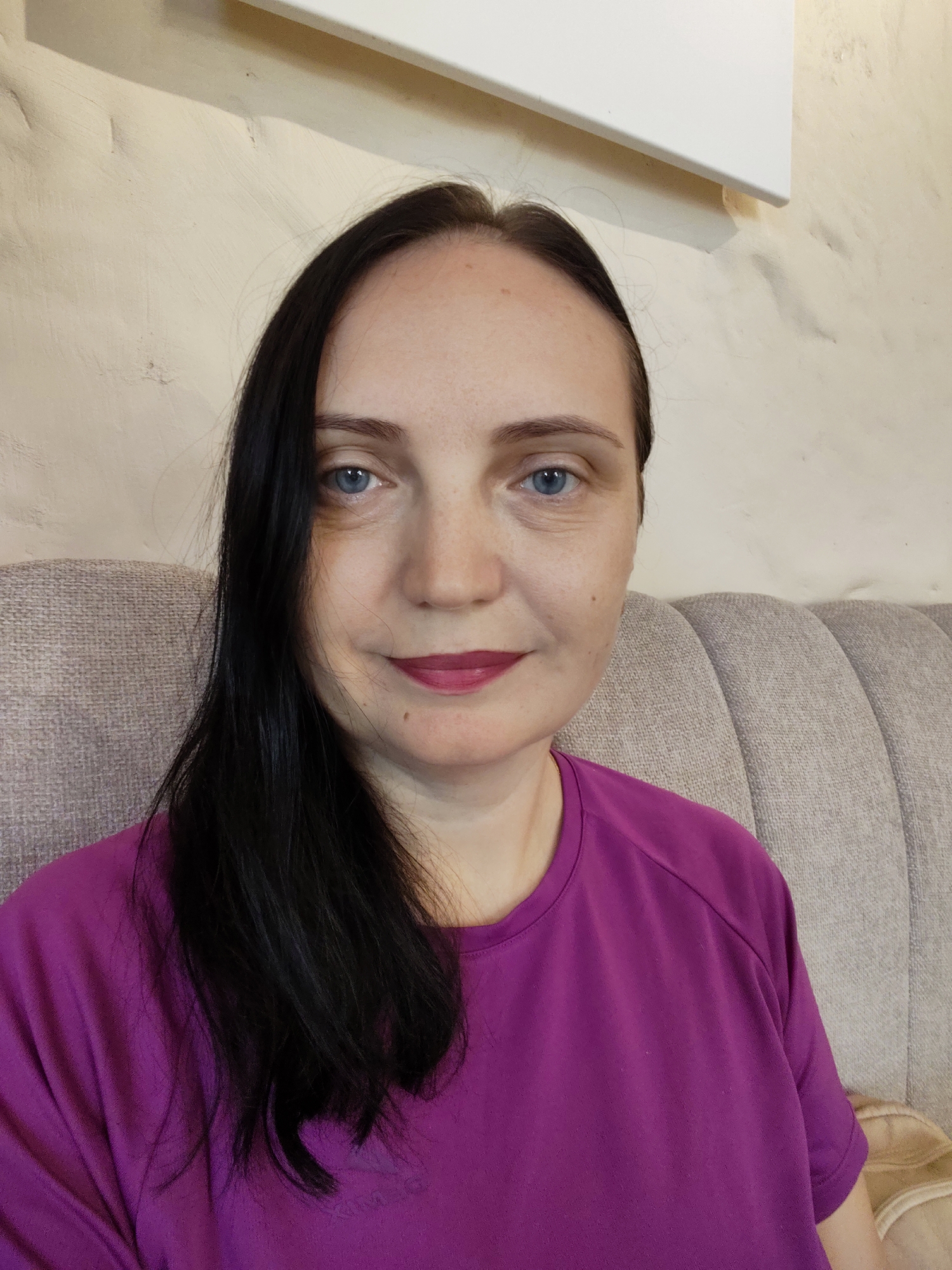 A medium shot taken indoors shows a woman with long, dark hair and blue eyes. She is wearing a bright purple t-shirt. Her face is turned slightly to the left, and she is looking directly at the camera. She has a light complexion and is wearing pink lipstick. The background is a light-colored wall with some texture, and a padded sofa with vertical ribs. The image appears to be a selfie, likely taken in a residential setting in Sochi, Russia, possibly her home. There are no other people or significant objects in the frame.