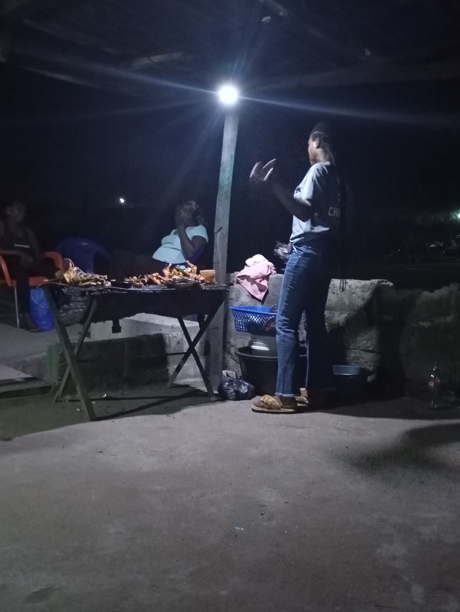 In the city of Shendam, Nigeria, at night, a group of people are gathered around a grill. The scene is dimly lit by a bright overhead light. In the foreground, a person in jeans and sandals stands with their hands gesturing. To their left, a table is laden with grilled meat. Two other individuals are visible in the background, one sitting and the other standing near the grill. The environment appears to be an outdoor, possibly informal, dining or food preparation area, with concrete flooring and some makeshift seating.