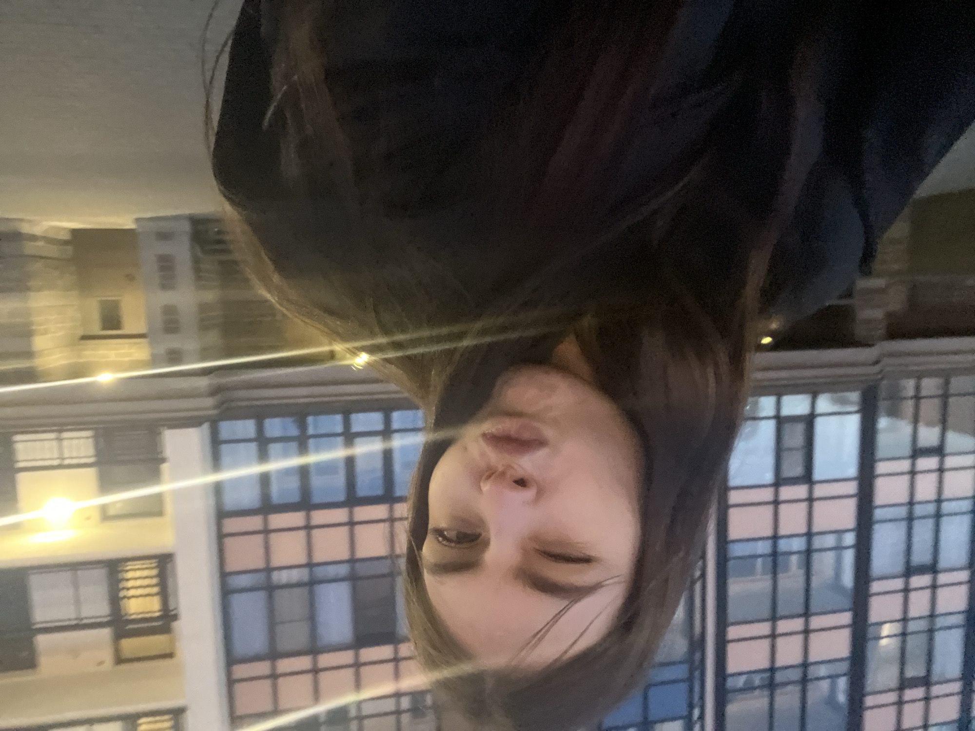 A young woman poses in front of a building in Vsevolozhsk, Russia. The building features a grid-like pattern of windows. The woman has dark hair, is winking one eye, and has slightly puckered lips. She is wearing a dark top. The image is rotated ninety degrees, and there are visible lights from nearby buildings.