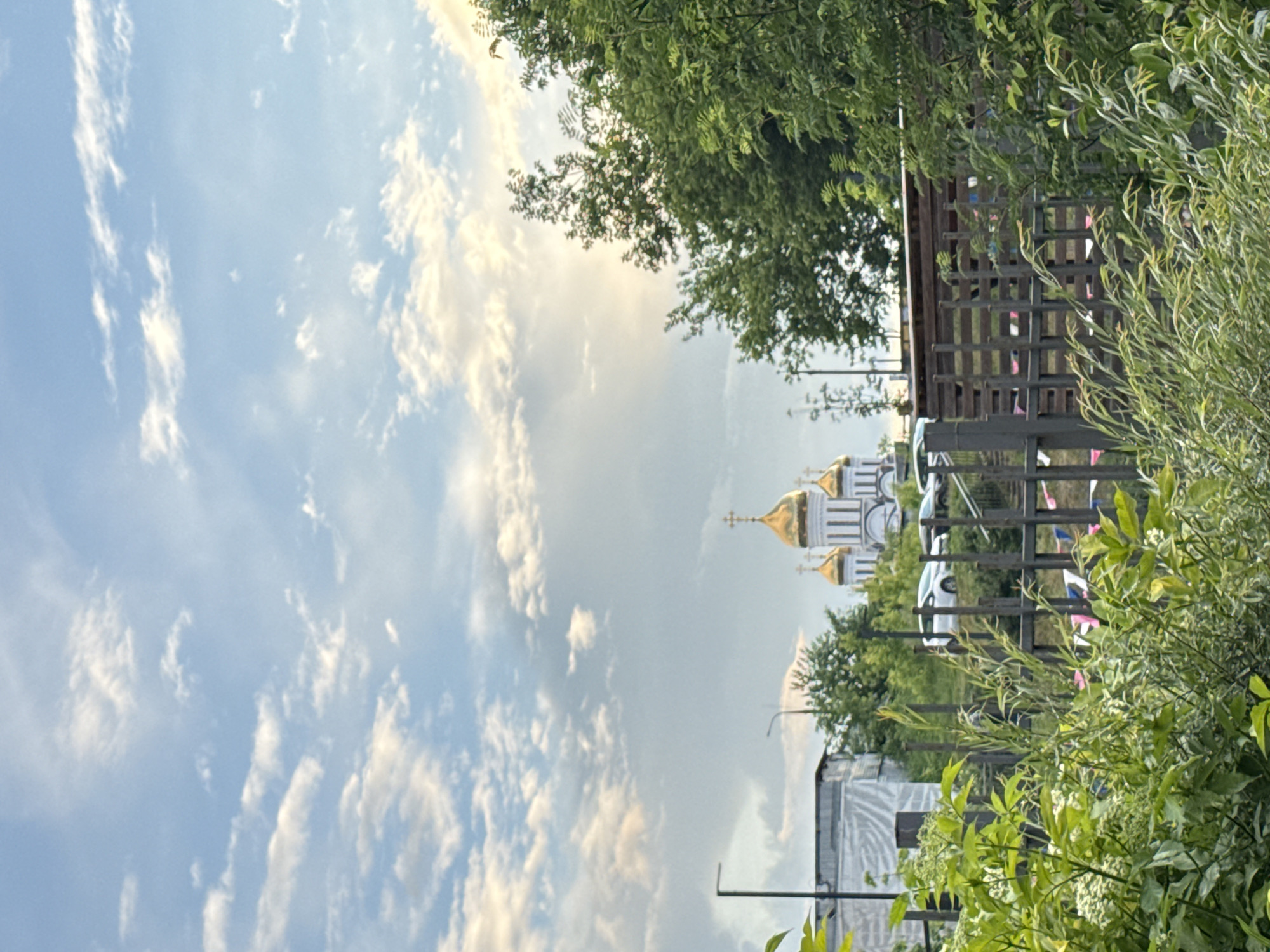 The image depicts an outdoor scene with a prominent Orthodox church in the background, characterized by its white facade and golden domes, suggesting a location in Moscow, Russia, given the architectural style and common presence of such churches in the city. In the foreground, dense green foliage and trees obscure the lower portion of the scene. A fence made of dark metal and wooden planks runs horizontally across the middle ground, partially separating the viewer from the background. Beyond the fence, two white cars are visible parked on a sloped area. To the left of the church, there is a low-rise building covered with white sheeting and a lamppost. The sky is partly cloudy, with a mix of white and grey clouds, and a soft, warm light suggests it may be late afternoon or early evening. There are no people visible in the image. The overall impression is one of a serene urban landscape blending nature, religious architecture, and everyday life.