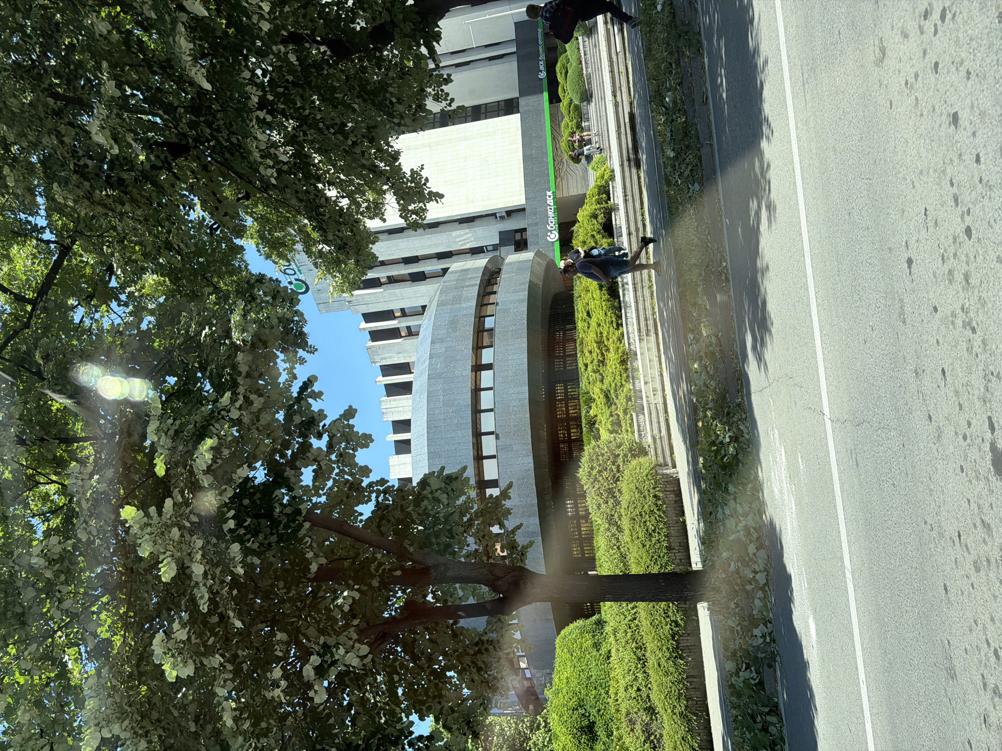 The image shows the DSK Bank building in Varna, Bulgaria. The building has a modern architectural design with a curved section in the foreground and a taller, angular section in the background. Lush green landscaping surrounds the building, with manicured bushes and trees. The sky is clear and blue, suggesting a sunny day.
Several people are visible in the image. In the middle ground, a man is walking past a series of steps leading up to the building. Further to the right, two people are seated on the steps, and another person is walking away from the camera. In the foreground, a person is walking to the left.
The perspective of the photo is from a slightly elevated position, possibly from a vehicle moving on a road that runs in front of the building. A white line is visible on the asphalt road in the foreground. The image is characterized by dappled sunlight and shadows cast by the trees, indicating a bright, outdoor setting.