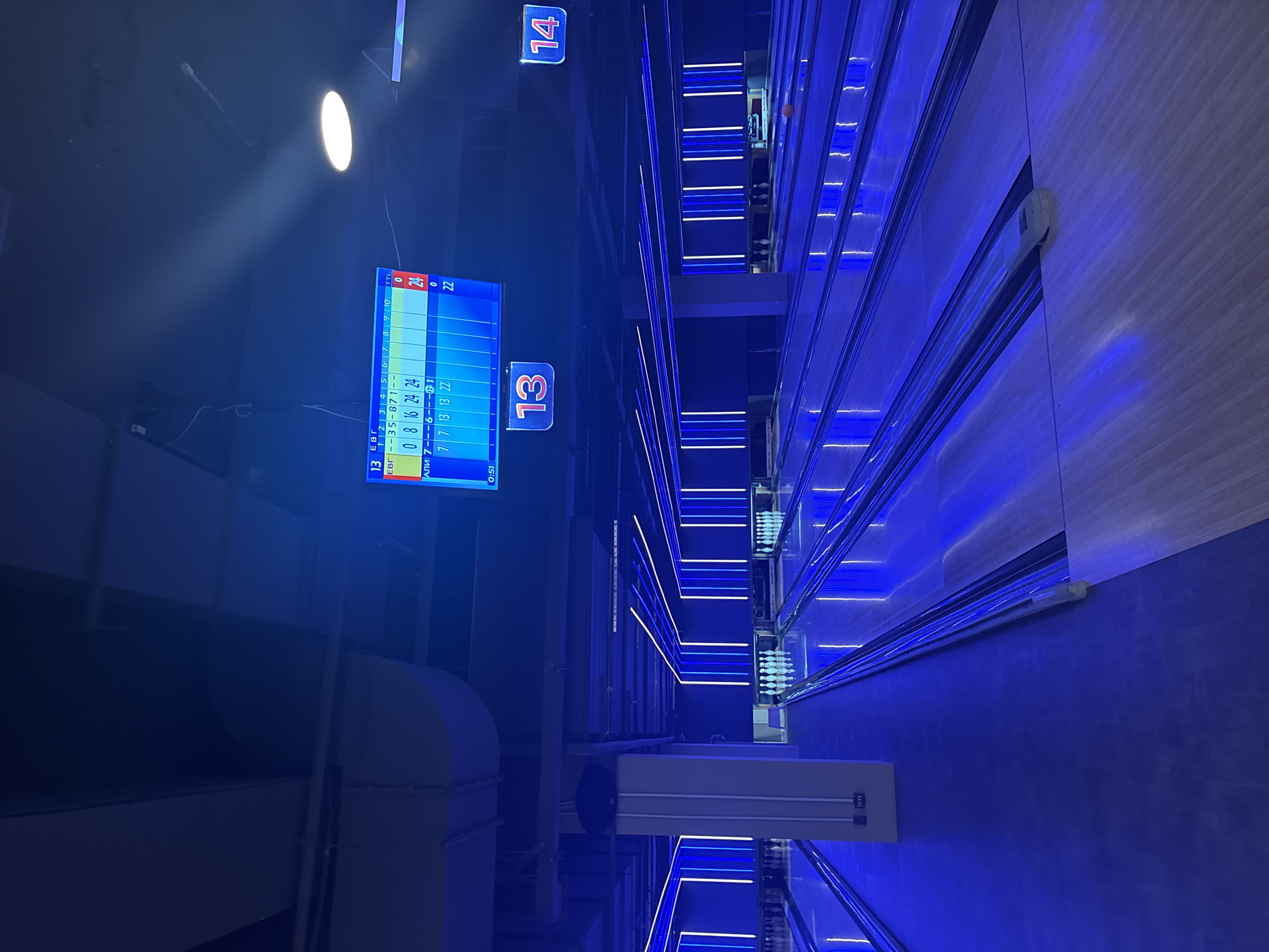 This is a photograph of an indoor bowling alley, illuminated by vibrant blue neon lights. The scene depicts multiple bowling lanes stretching into the distance, with pins set up at the end of each. A bowling ball is visible on one of the lanes towards the right side of the image. Overhead, a digital scoreboard displays scores for a game, showing frame numbers and individual frame scores. Two lane numbers, "13" and "14," are prominently displayed in illuminated red frames. The overall environment suggests a modern entertainment venue. The image is a photograph taken in Saint Petersburg, Russia.