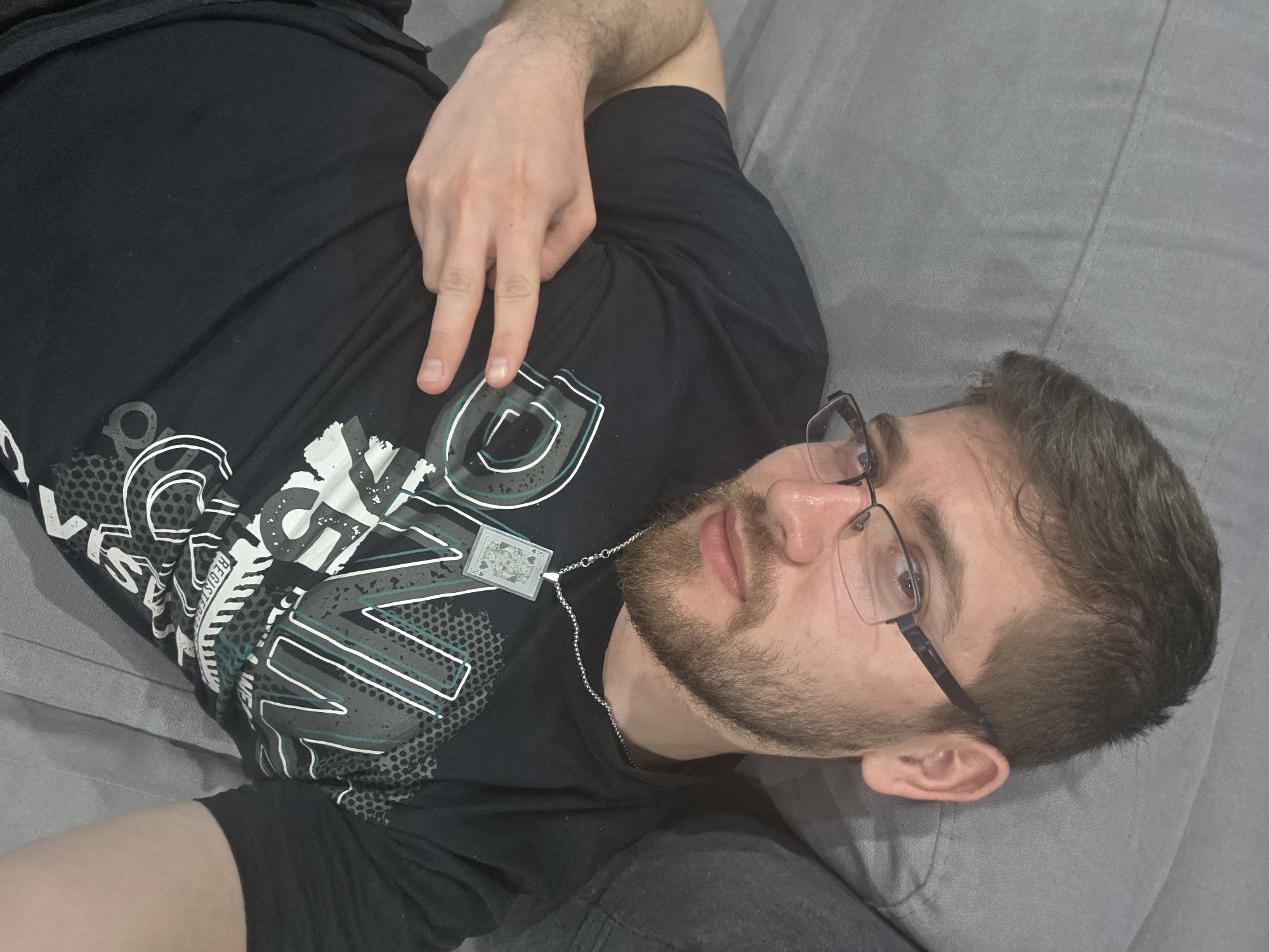 A man is lying on a grey sofa, facing upwards. He is wearing glasses and a black t-shirt with a white and grey graphic design and a playing card pendant. He is making a peace sign with his right hand. The setting is indoors and appears to be a residential space. There's no information that indicates the location in the city of Yagodovo, Bulgaria.