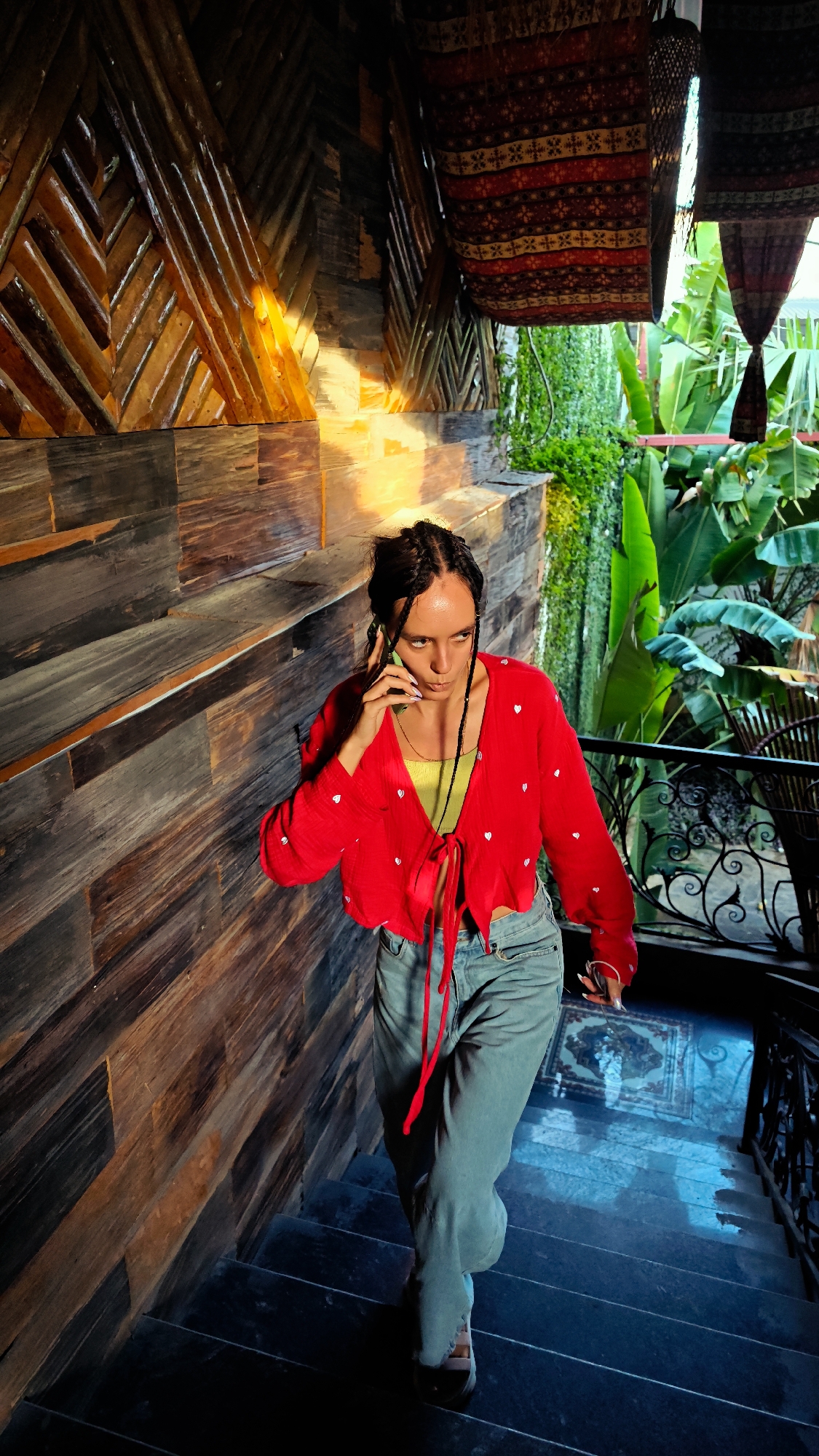 The image captures a female individual descending a dark staircase within an architectural space in Trung Tam Ward, Vietnam. The subject, a young adult with dark braided hair, wears a red cropped cardigan adorned with small white hearts over a lime green top, light blue jeans, and light-colored shoes. She is holding a light green smartphone to her right ear, appearing to be in conversation, with an intense, upward gaze. To her left, a wooden wall displays a mix of horizontal planks and a diagonal, chevron-like pattern, illuminated by a warm, golden light source. To her right, a decorative wrought-iron railing follows the curve of the stairs. Beyond the railing, dense tropical foliage, including large banana-like leaves, is visible under natural light. Overhead, several patterned textiles with traditional designs in red, yellow, and dark hues hang decoratively. The dark stairs transition to a visible section of blue and white patterned tiled flooring at the bottom, which is distinctly illuminated by a blue light.