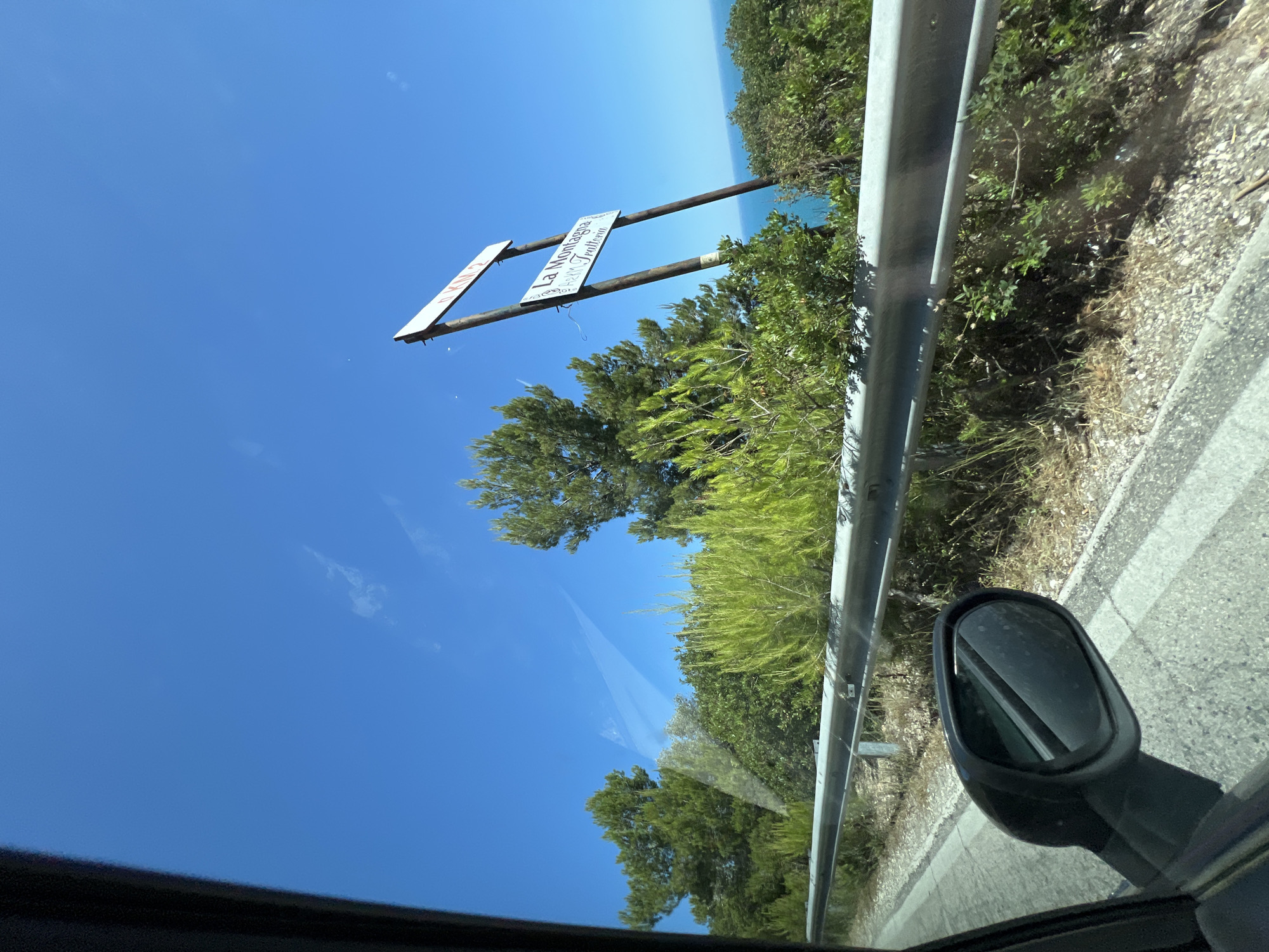 The image shows a roadside view on a clear, sunny day with a bright blue sky. A metal guardrail runs along the side of the road, separating it from a slope covered in dry grass, gravel, and various green shrubs and trees. In the background, a sign with text is mounted on two wooden posts. The upper part of the sign is white with red lettering that appears to say "D. KM. 2." The lower sign is also white with black lettering, reading "La Montagna" and below that, "Trattoria." Beyond the vegetation and the sign, a glimpse of the blue sea can be seen. The foreground includes the interior of a car, with a portion of the car's window and a side mirror visible on the left. The scene appears to be located on a coastal road, potentially near Mattinata, Italy, given the presence of "La Montagna Trattoria," which is a known establishment in the area.