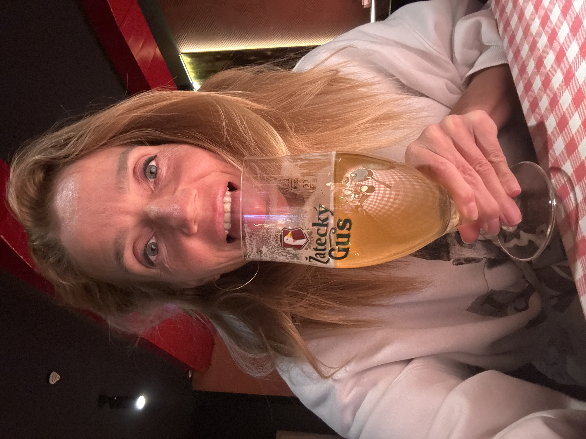 A person with long, blonde hair is tilting their head towards the camera, holding a glass of beer with a "Žatecký Gus" logo on it. They are smiling and the glass is partially obscuring their face. They are wearing a white shirt and a table with a red and white checkered tablecloth is visible to the right. The background shows a dark interior with red and black walls, and some decorative lighting. There is no definitive evidence indicating the location is Tashkent, Uzbekistan.