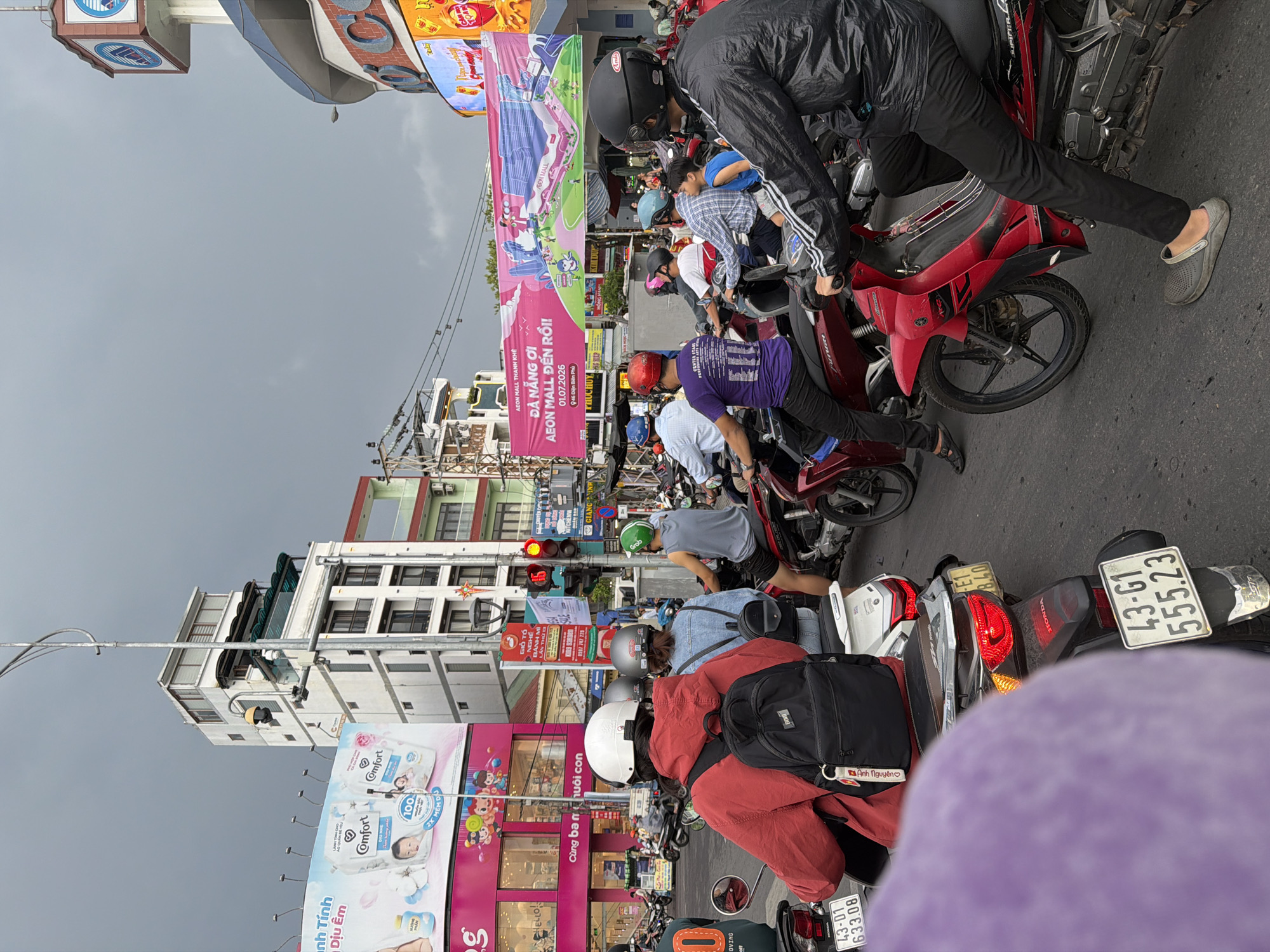 This is a photograph taken in Đà Nẵng, Vietnam, depicting a bustling street scene with heavy motorcycle traffic. The camera angle is from behind and slightly above, looking down at the road. The sky is overcast, suggesting a cloudy or possibly rainy day.
The primary subjects are people riding and standing around a multitude of motorcycles. Many individuals are wearing helmets, with some in casual clothing and others in jackets. The motorcycles are packed closely together, indicating a traffic jam or a crowded marketplace.
Prominently visible are several large banners and billboards. One banner, in pink, advertises "AEON MALL THANH KHÊ" and announces its opening on "01.07.2026" at "946 Điện Biên Phủ". Another billboard features an advertisement for "Comfort" fabric softener, showing images of babies and product packaging with text in Vietnamese. Other signs in Vietnamese are also visible on buildings and stalls.
The setting appears to be an urban street with multi-story buildings lining the road. Traffic lights are visible, showing a red light, further confirming the presence of traffic congestion. The overall impression is one of a busy, everyday scene in a Vietnamese city, characterized by the ubiquitous presence of motorcycles and commercial advertising. The mood is one of energetic, albeit perhaps frustrating, urban life.