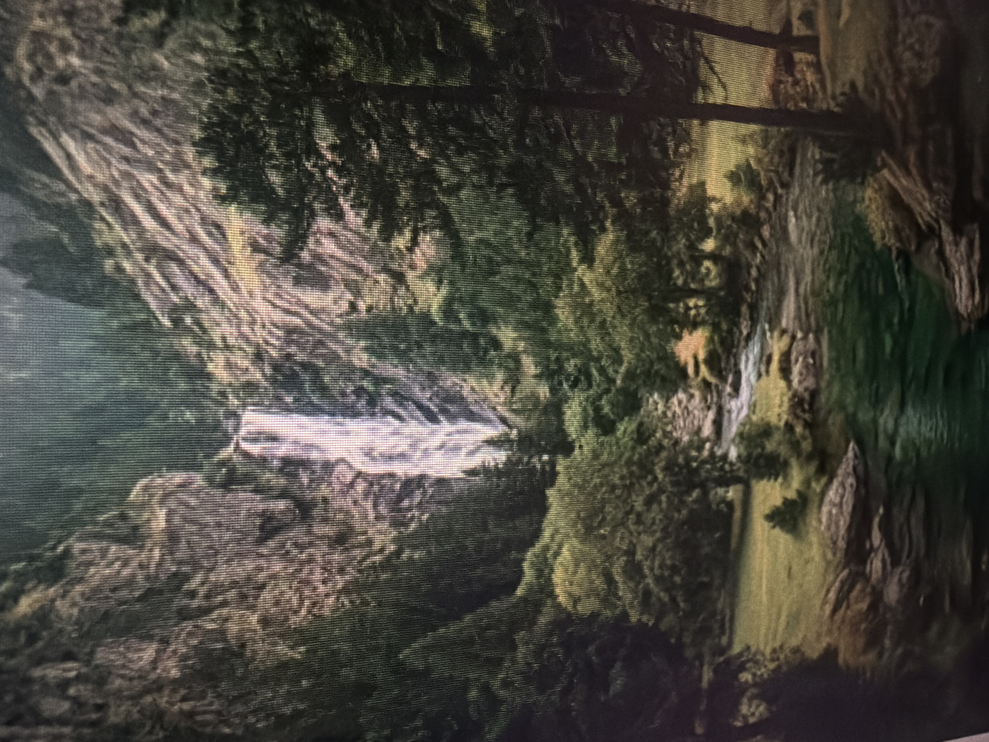 The image depicts a scenic natural landscape featuring a tall waterfall cascading down a rocky cliff face. The waterfall is the central element, surrounded by dense green foliage and trees. A river flows through a grassy clearing in the foreground, with rocks and smaller trees along its banks. The overall scene suggests a serene and wild environment, likely in a mountainous or forested region. The location in Baan Mae Nam, Thailand, is not identifiable from the visual content of the image itself, as there are no specific landmarks, cultural elements, or geographical markers that indicate this particular place.