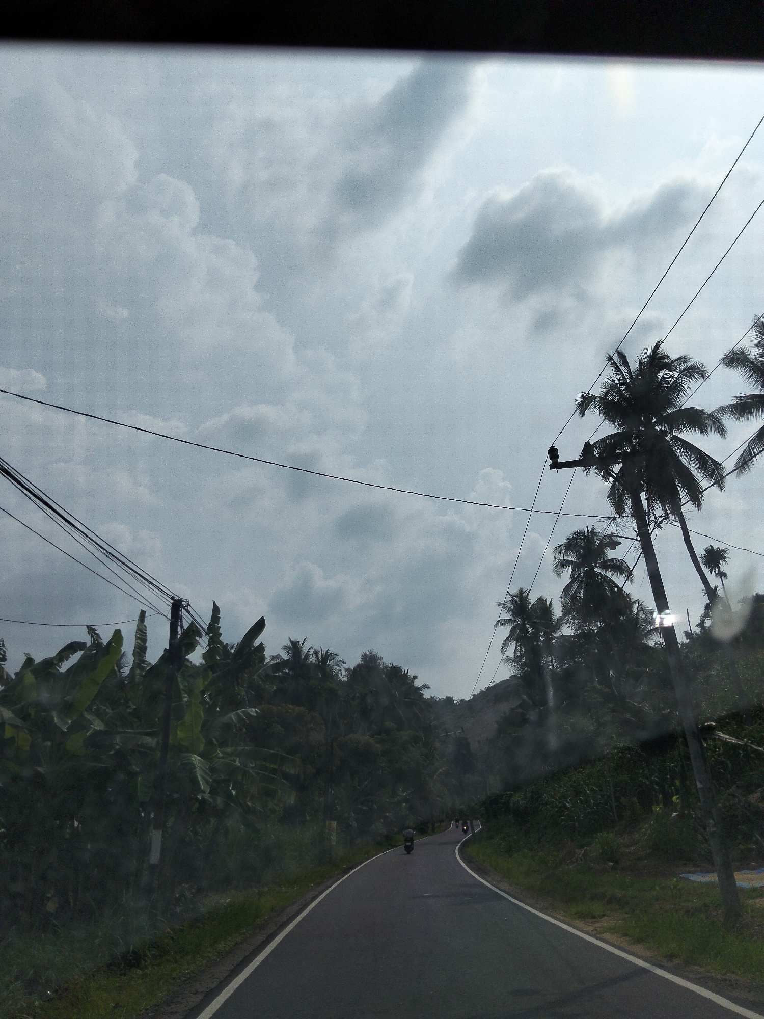 The image shows a view from a vehicle driving along a road in Luengbaro, Indonesia. The sky is overcast with dramatic, grey clouds, suggesting it may be an afternoon or late morning. Tall palm trees and lush green vegetation, including banana trees, line the sides of the road, indicating a tropical environment. Power lines crisscross the sky, supported by utility poles. The road itself is paved and curves to the left, leading into the distance. In the far distance, a few motorcycles can be seen on the road, suggesting a mode of transportation common in the area. The overall mood is somewhat subdued due to the cloudy weather, but the vibrant greenery offers a sense of life and natural beauty. There is no visible text in the image.
