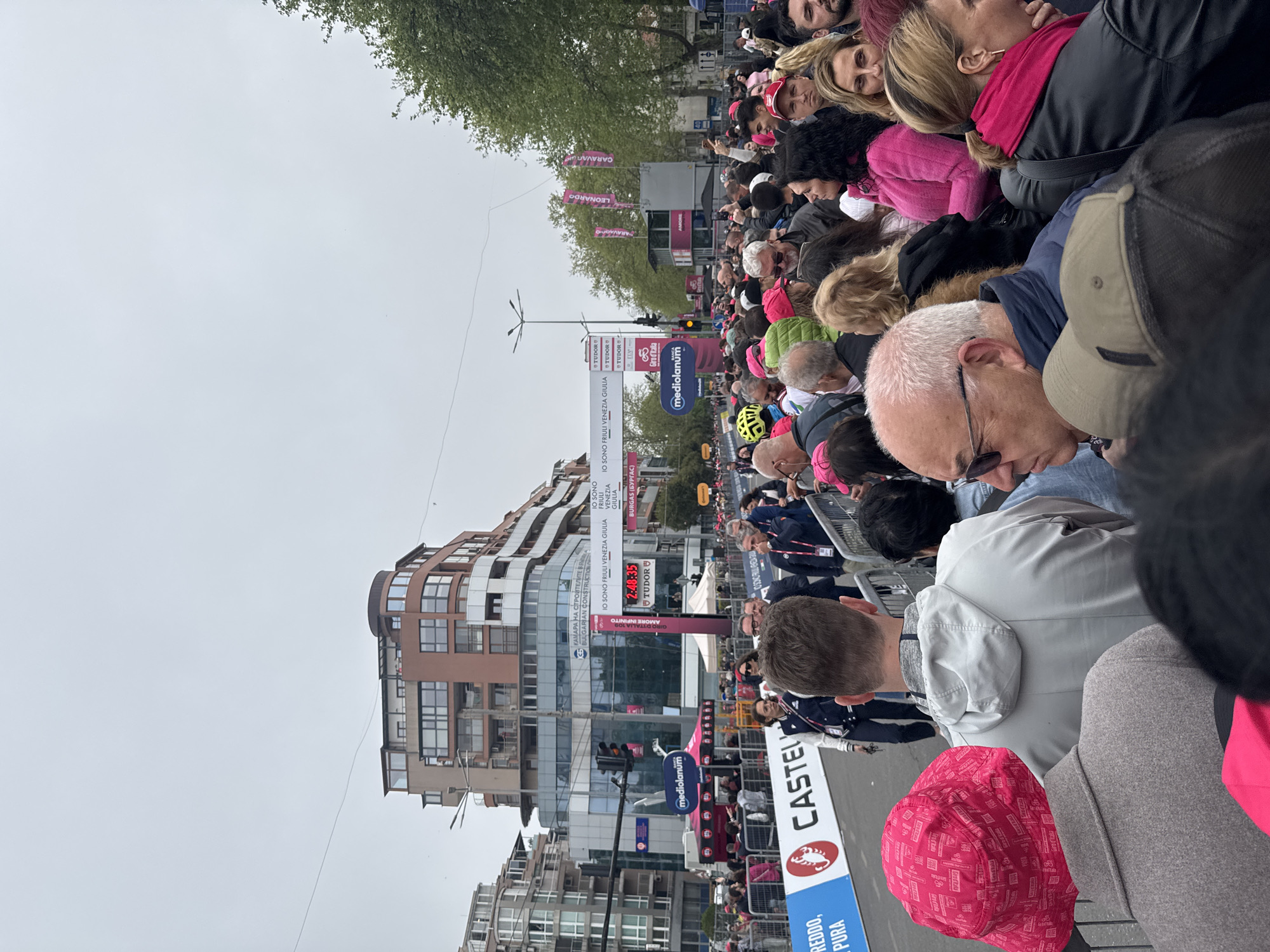 The image shows a crowd of people gathered to watch the Giro d'Italia in Burgas, Bulgaria. A clock displays the time of the race as 2:40:35. Banners with the names of Italian regions, "IO SONO FRIULI VENEZIA GIULIA," and the sponsors "TUDOR" and "mediolanum" are visible. A sign "CASTEL" and "REDDO, PURA" is visible in the foreground. Several buildings and a traffic light are in the background, along with overhead light poles. The crowd is densely packed, with many wearing pink, and is separated from the road by metal barriers.