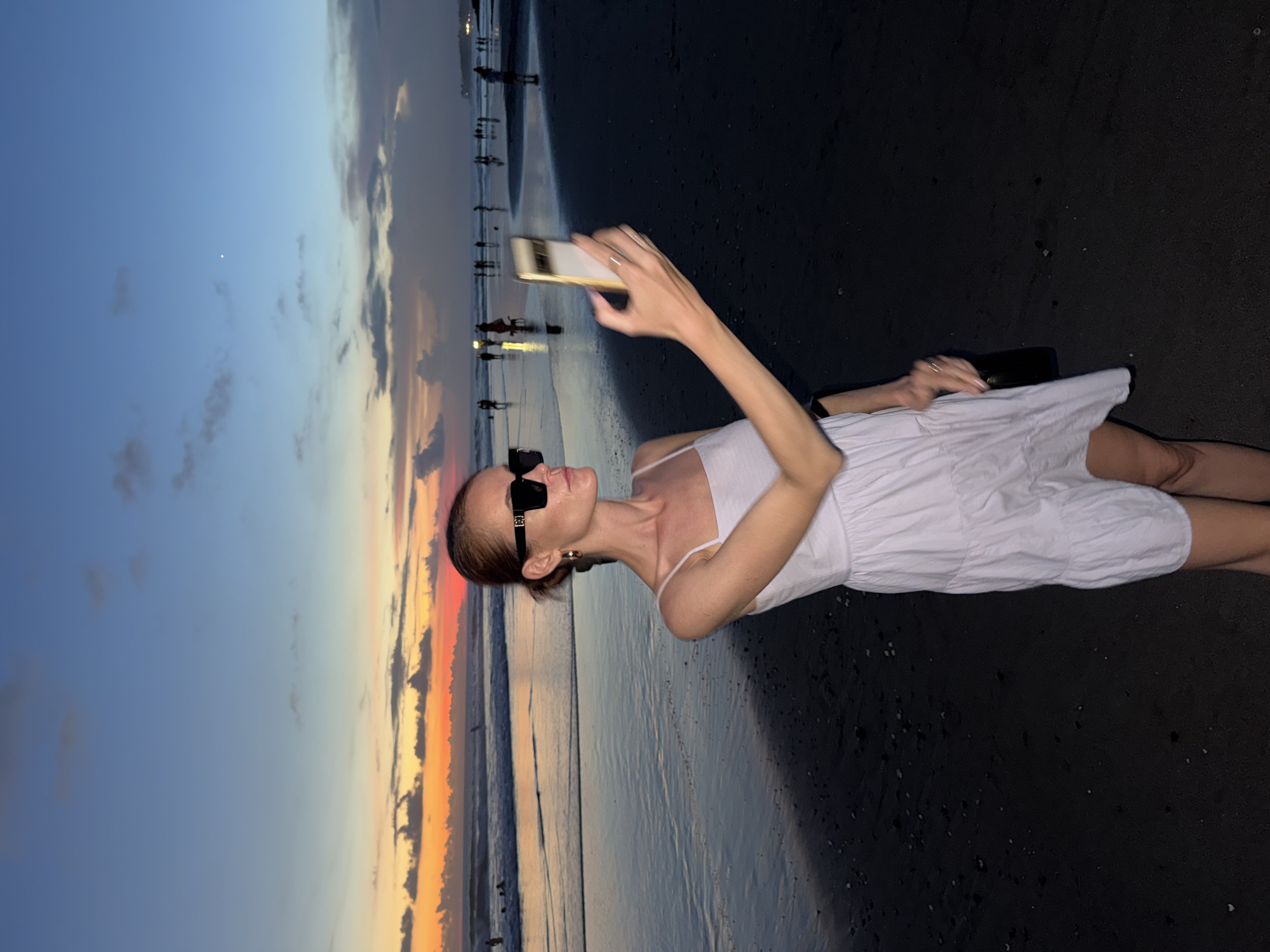 A woman is standing on a dark sand beach at sunset, seemingly taking a photo of the view with her smartphone. She is wearing a white sundress and dark sunglasses. People are silhouetted in the background along the water's edge. The sky is a mix of blues and oranges due to the setting sun. The location is North Kuta, Indonesia, based on the beach's dark sand, which is typical of the area.