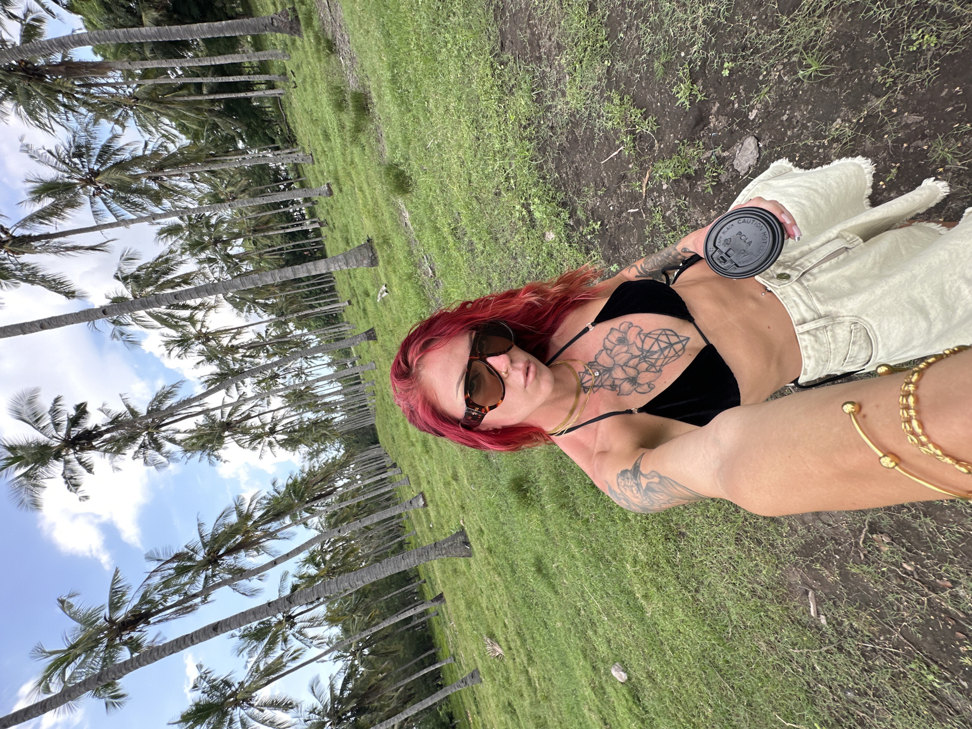 The image is a selfie taken by a person with vibrant red hair and dark sunglasses, set against an outdoor, tropical backdrop. The individual wears a black top, possibly a bikini or bralette, and light-colored pants or a skirt with a fringed hem. Several tattoos are visible on their arms and chest, including an intricate geometric or floral pattern on the upper chest and a bird design on the bicep. They are adorned with gold chain necklaces, a nose ring, and gold bangles on their wrist.
In their hand, they hold a black disposable coffee cup with a lid. Visible text on the lid includes "CAUTION HOT," "PCLA," and a recycling symbol.
The background features a dense grove of numerous tall palm trees with green fronds, extending into the distance under a clear blue sky with scattered white clouds. The ground is covered with green grass interspersed with patches of dark soil. The scene suggests a bright, sunny daytime, evoking a relaxed, possibly vacation-like atmosphere in a warm climate.