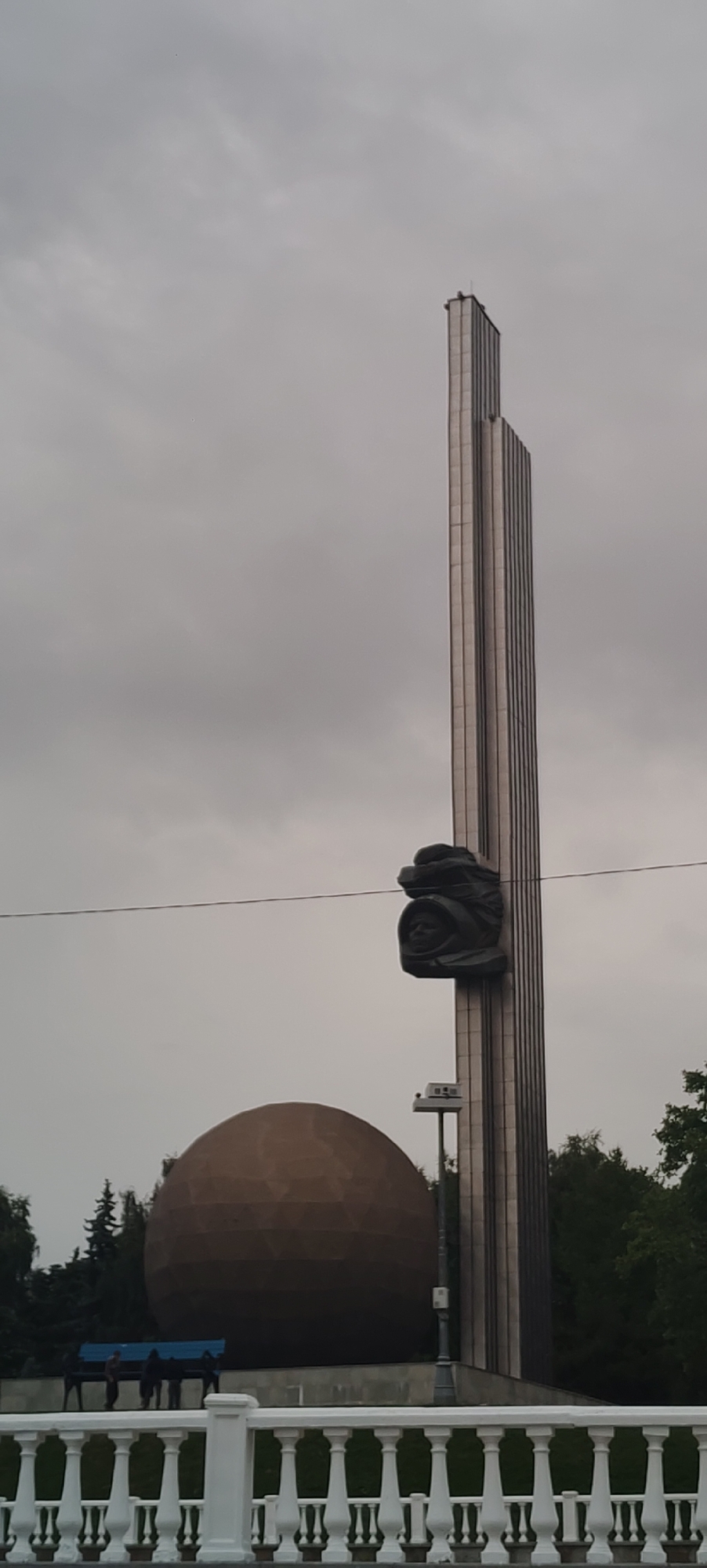 The image depicts a monument in Kaluga, Russia, likely the "Tsiolkovsky Monument" or a similar space-themed memorial, given its features. The central element is a tall, fluted metallic obelisk reaching towards a heavily overcast sky. Mounted on the obelisk at mid-height is a dark bronze sculpture of a human head wearing a helmet, suggesting a connection to space exploration. To the left of the obelisk, a large, geodesic dome-like structure, colored a muted brown, stands prominently. This dome might represent a planet or a celestial body. In the foreground, a white balustrade with vertical spindles separates the viewer from a grassy area and a paved walkway. Behind the balustrade, several dark figures are visible, appearing to be standing or sitting on a bench in front of the dome. Trees, likely deciduous and coniferous, form a backdrop to the monument and dome. The overall atmosphere is somber due to the cloudy sky.