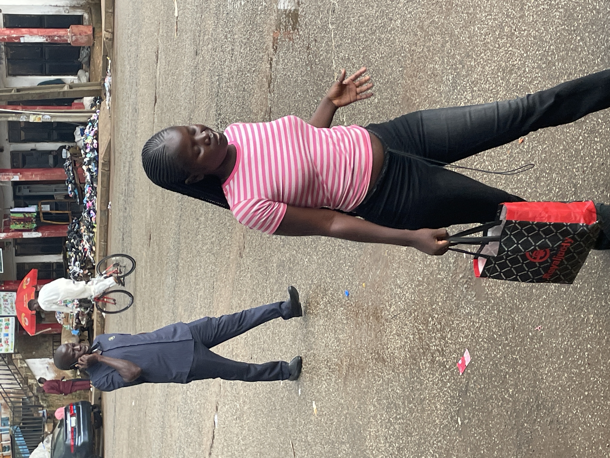 In Bukuru, Nigeria, a man in a dark blue pinstripe shirt and dark trousers is on the phone, walking on a wet asphalt surface. Behind him, a person in white is riding a red bicycle. In the foreground, a young woman with dark skin and braided hair is walking towards the right, wearing a pink and white striped t-shirt and dark jeans. She is carrying a black and red shopping bag. The background shows shops with goods displayed outside, and some vehicles, including a dark blue car. The ground is littered with small pieces of paper and debris. The overall scene depicts a busy street with everyday activities.