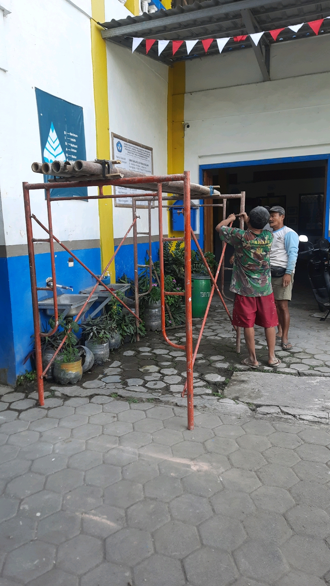 Two men are seen in front of a building with blue and yellow accents, located in Nanggulan, Indonesia. The building has a sign that reads "SMA SANJAYA XXI NANGGULAN". Scaffolding is erected in front of the building, and one of the men, wearing a camouflage shirt and red shorts, is interacting with the scaffolding. The other man, in a light blue and white shirt and shorts, stands nearby. The ground is paved with hexagonal stones. There are plants in pots and a green barrel visible. A motorcycle is parked to the right of the frame.