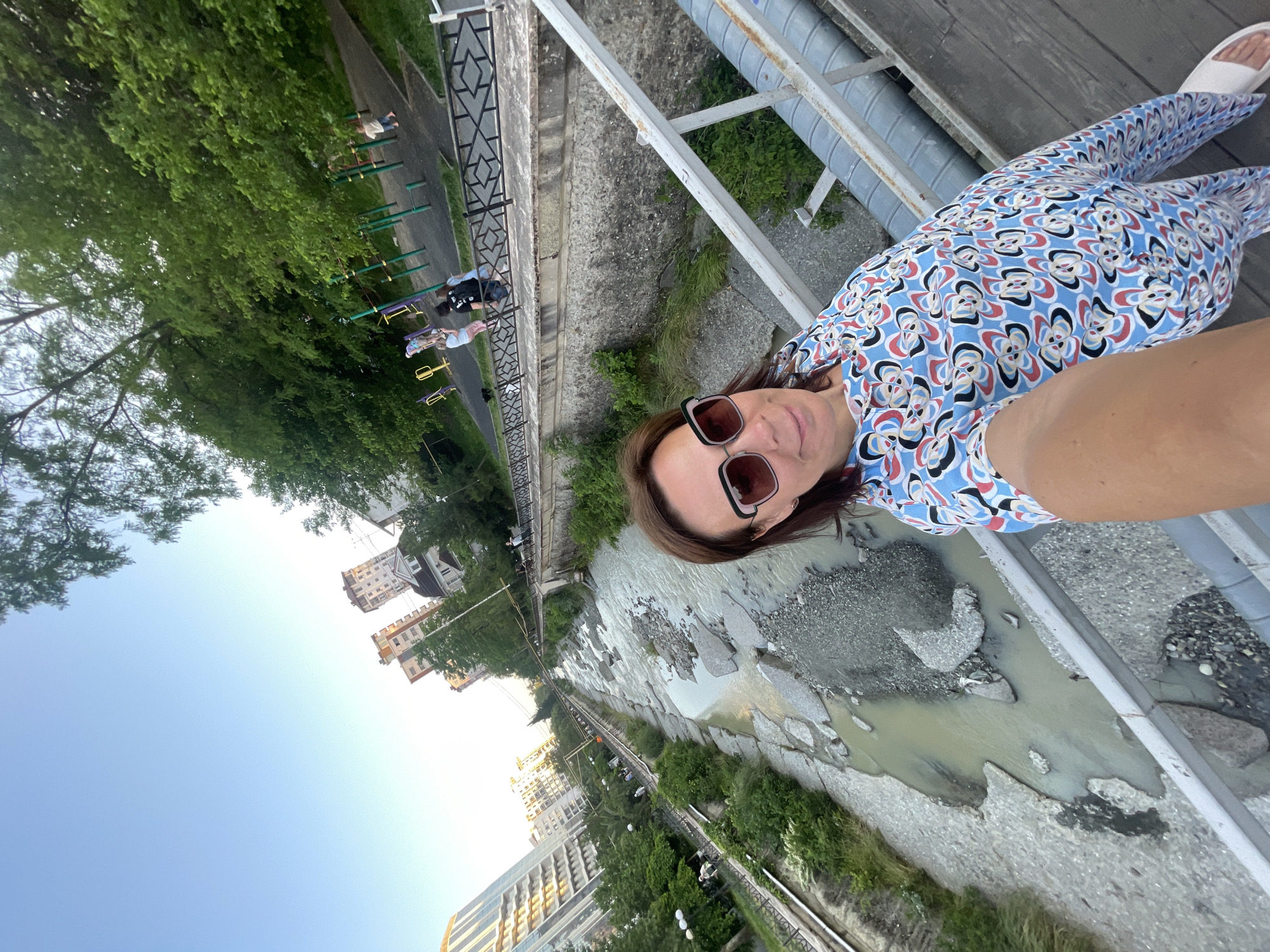 An adult woman is depicted in a high-angle selfie taken from a pedestrian bridge over an urban river in Sochi, Russia. The woman, positioned in the foreground and slightly right of center, has brown hair and wears dark-framed sunglasses. She is smiling faintly and dressed in a light blue jumpsuit or dress patterned with stylized white, red, and black floral or bow-like shapes. Her right arm, holding the camera, is partially visible at the bottom of the frame. She wears white flip-flops and stands on wooden planks, which form the surface of the bridge. Metal railings and utility pipes run along the bridge structure to her right.
The river below is shallow and wide, with murky, light brown water revealing a bed predominantly covered in exposed grey rocks and gravel. On the left bank of the river, an embankment features a paved walkway lined with a black metal fence and green trees. Several pedestrians are visible walking along this path, and multi-story modern residential buildings, including one with a reflective facade, rise in the background. Streetlights are also present along the walkway.
On the right bank, directly behind the woman, a concrete retaining wall with some natural vegetation supports an elevated area. Above this wall is a black metal fence with a diamond-pattern design. Beyond the fence lies a children's playground with colorful equipment, including yellow, green, and purple structures. Several children and adults are present in the playground, with one child barefoot. Lush green trees densely populate the area behind the playground, and further back, additional residential buildings, including one with a distinctive pitched roof and multiple gables, are visible, alongside other tall apartment blocks. The sky is clear and light blue, indicating daylight, likely late afternoon or early evening.