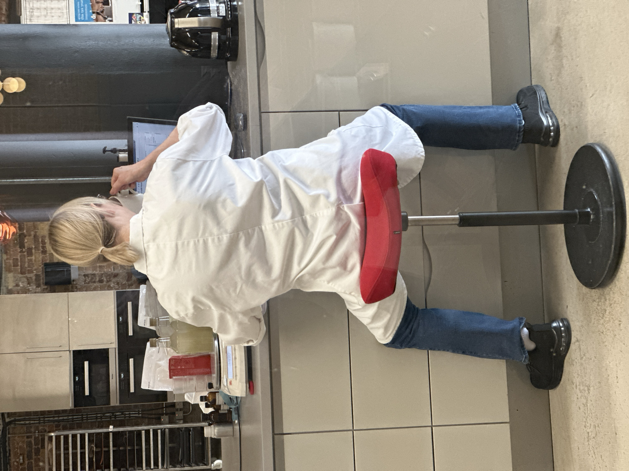 The scene depicts an adult individual, observed from behind, seated at a grey countertop in what appears to be a commercial or industrial test kitchen or laboratory facility located in London, United Kingdom. The individual has blonde hair tied in a ponytail and wears a white lab coat over blue jeans and black athletic footwear. They are positioned on a red, height-adjustable saddle stool. The individual's right hand holds a white mug to their face, while their attention is directed towards a laptop computer displaying data or a spreadsheet. To the left of the laptop, the countertop holds a white digital scale indicating "-4.90," flanked by multiple items including small silver and dark amber glass bottles, a red plastic container, clear plastic bowls, two corked glass bottles containing a pale yellow liquid, and white bags of ingredients. A black electric kettle and a small black pump dispenser are situated on the counter to the right of the laptop. The background features light grey, flat-panel kitchen or lab cabinetry, including integrated Miele ovens with digital displays. To the far left, an industrial metal shelving unit on wheels is visible against an exposed red brick wall. Prominent dark grey structural columns are present towards the right, behind which a partially obscured poster with the text "spring into action this tax year" is visible, strongly suggesting a United Kingdom location. The floor is a light, speckled concrete.