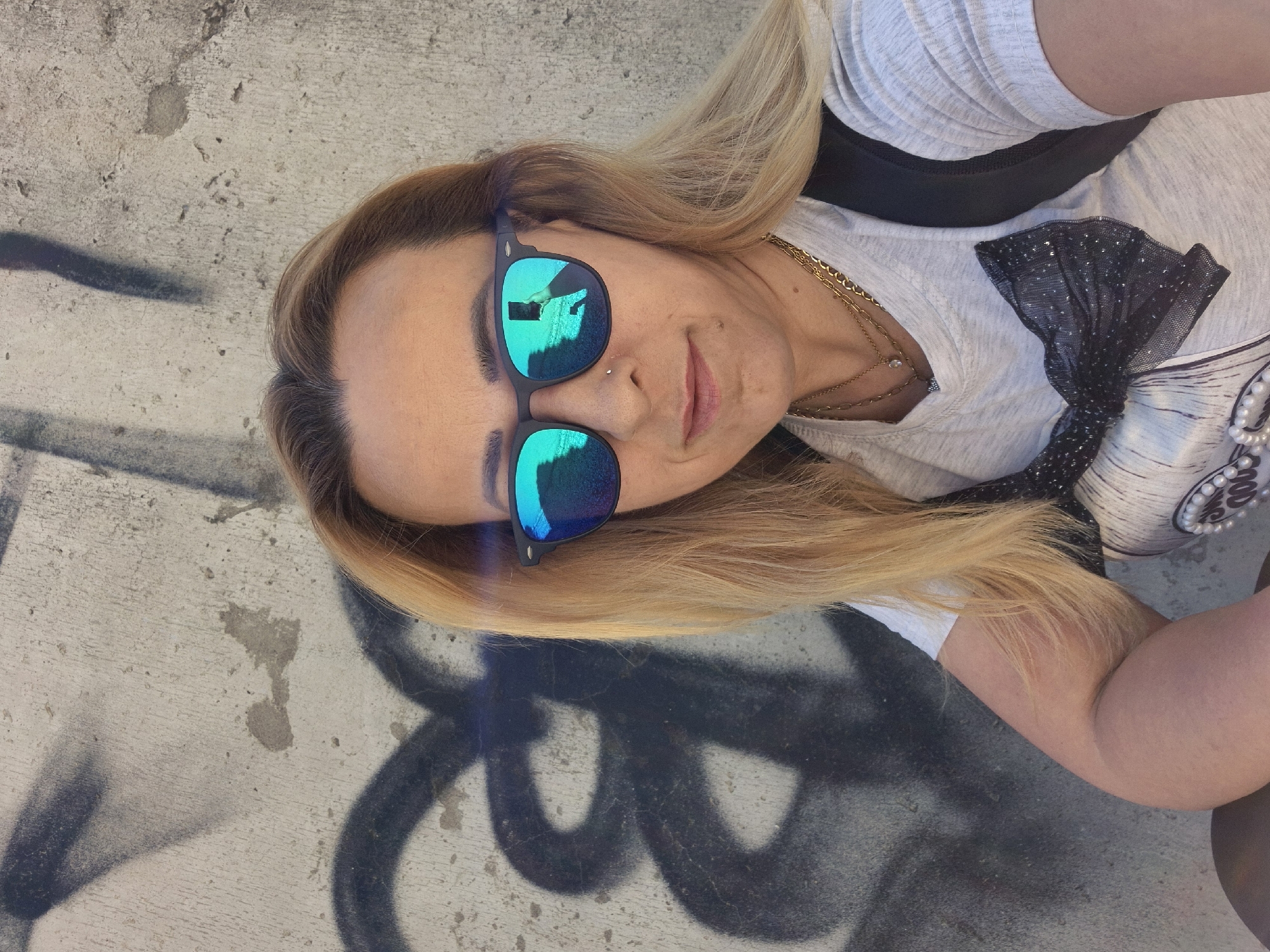 A medium shot captures a person from the chest up, posing for a selfie outdoors against a concrete wall covered in black spray-painted graffiti. The person, with blonde hair and wearing sunglasses with blue lenses, has a nose ring and a small mole on their right cheek. They are dressed in a light-colored t-shirt with a black sequined bow graphic on the front, accessorized with two gold necklaces. A black backpack strap is visible over their right shoulder. The sunglasses reflect the person holding a phone, suggesting they are taking the photo. The sunlight casts shadows on the wall. The location is not specified beyond "Unknown, Unknown."