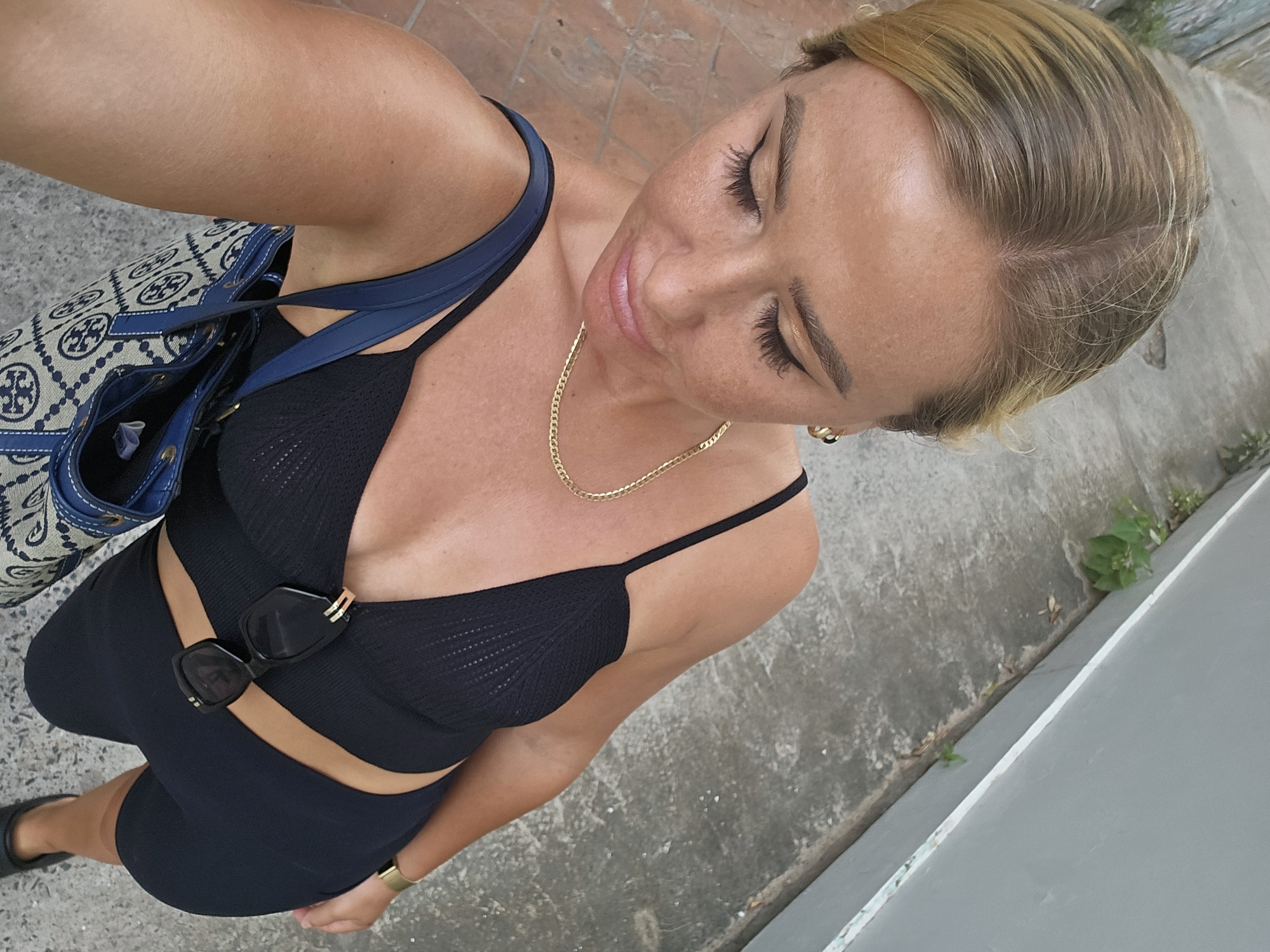 In Phu Thuy Ward, Vietnam, a woman with blonde hair pulled back stands on a sidewalk. She is wearing a black, ribbed tank top with a cutout and matching shorts, a gold chain necklace, and sunglasses clipped to her top. A blue and white patterned tote bag hangs from her left shoulder. The ground is paved with concrete and reddish tiles, and a grey wall runs along the right side of the scene, with some small plants growing at its base. The lighting suggests it is daytime.