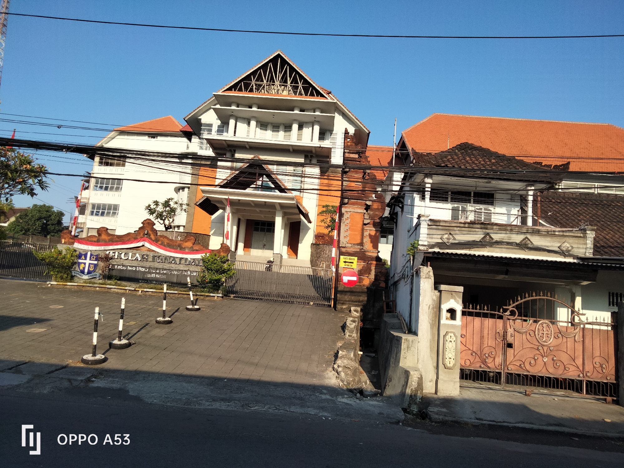 The image depicts a street scene in Denpasar, Indonesia, on a clear day with a bright blue sky. The foreground shows a paved road with some cracks and a concrete curb. Several black and white striped bollards are lined up on the right side of the road, leading towards an entrance.
Dominating the midground and background are several buildings. On the left, a white building with orange accents and a red tile roof is visible, with a sign that reads "SEKOLAH TUNA DAI" and an address "Jl. Kebo Iwa Utara No. 8 Denpasar Bali 80115". The building has multiple levels and large windows. Further to the right, another building with a prominent orange tile roof and white walls is seen, with a more traditional Balinese architectural style featuring intricate brickwork. Next to this, a smaller, white, two-story building with a dark tiled roof is partially visible. In front of the Sekolah Tuna Dai, a metal gate with a decorative pattern is visible. A yellow sign with red text and a circular red symbol is attached to a pillar near the gate.
The environment is characterized by overhead electrical wires crisscrossing the sky. Some trees are present, including a leafy green tree on the far left and a smaller tree with sparse foliage in front of the brick building. The overall impression is that of a street in a residential or institutional area of Denpasar.