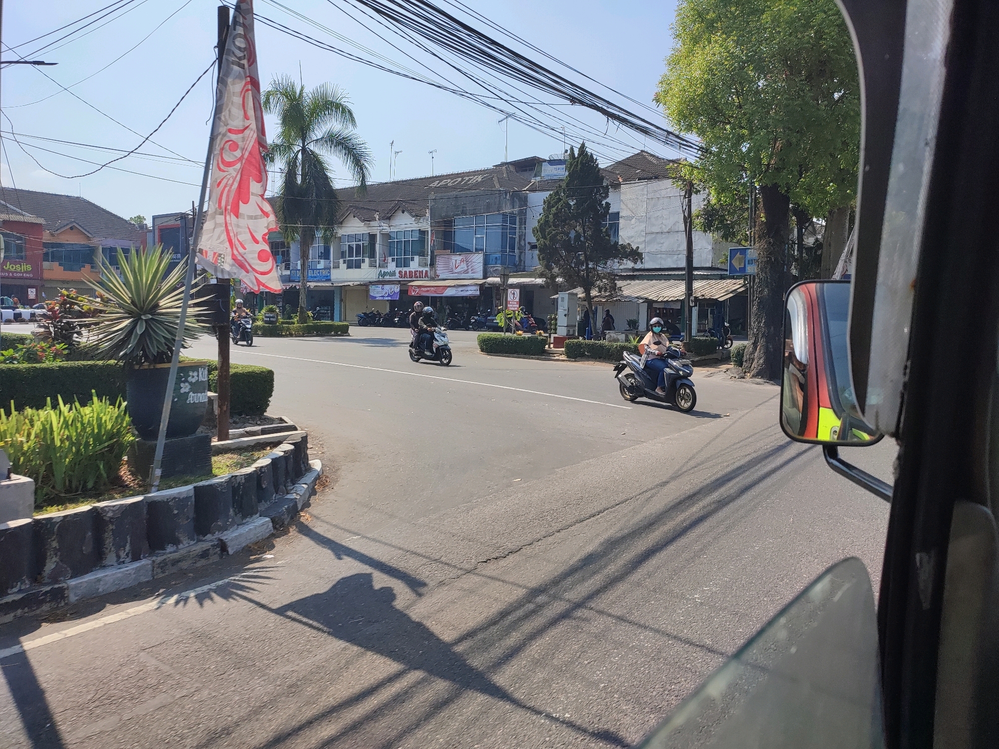 The image depicts a street scene in Magelang, Indonesia, likely near a commercial area. In the foreground, a portion of a vehicle, possibly a motorcycle or a car, is visible on the right side. A side mirror reflects the scene, showing a person wearing a helmet. The road is paved, with some cracks and shadows cast by overhead wires and trees.
On the left side of the road, there is a planter with green foliage and a decorative stone border. A flag with red and white patterns is attached to a pole. Beyond the planter, several buildings with a light-colored facade line the street. Some of these buildings appear to be shops or businesses, indicated by signage such as "Josilis" and "Apotek SABENA." A blue sign with white text is also visible.
Several individuals are seen riding motorcycles on the road, moving in different directions. One person on a motorcycle in the foreground is wearing a light-colored shirt and a helmet. Another motorcycle with two riders is further down the street. Palm trees and other leafy trees are present, indicating a tropical or subtropical climate. The sky is clear and blue, suggesting a sunny day. The overall impression is a typical urban street in Indonesia, with the usual elements of traffic, commerce, and vegetation.