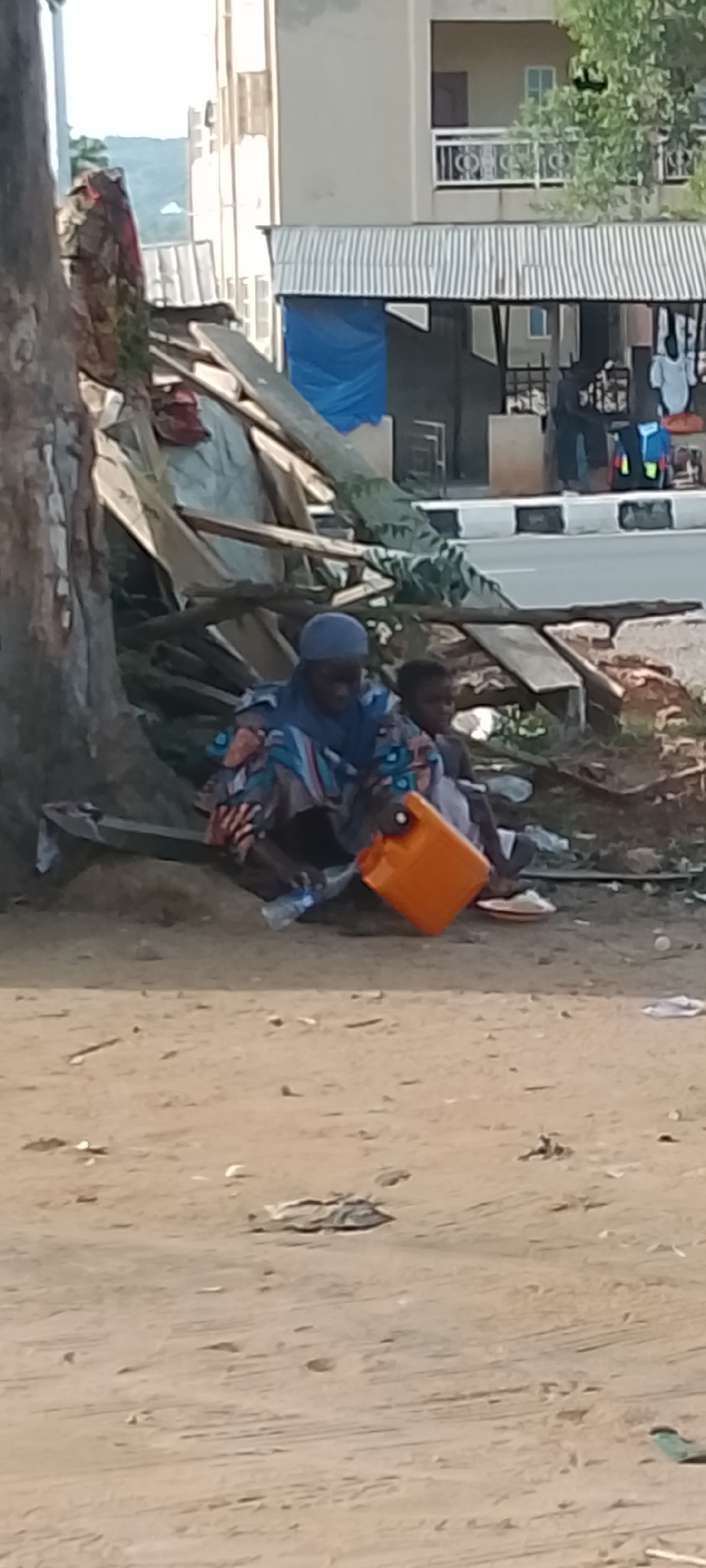In Adankolo, Nigeria, a woman and child are sitting on the ground next to a tree trunk and debris pile. The woman is wearing a head covering and patterned clothing, holding an orange container. A child sits beside her. They appear to be adjacent to a road with black and white painted curbs. A building with a blue tarp and clothing is visible in the background. The ground is dirt.