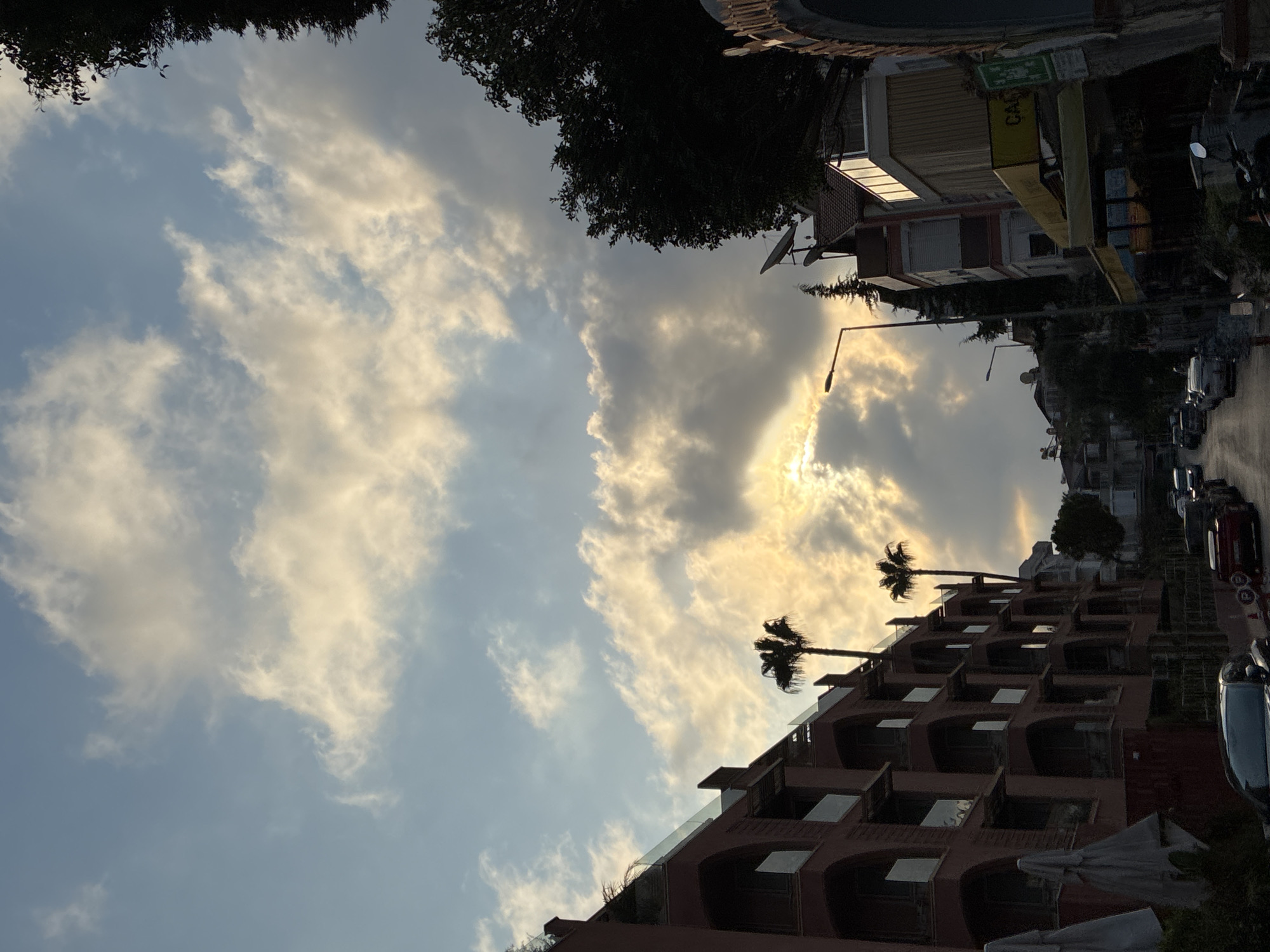 The image depicts a street scene in Alanya, Turkey, with a prominent building on the left and a street lined with parked cars and businesses. The sky above is filled with dramatic clouds, illuminated by the setting or rising sun, which peeks through the clouds, casting a warm glow.
The building on the left is several stories high, with a reddish-pink facade and arched balconies. Palm trees are visible in front of it, silhouetted against the bright sky. On the right side of the street, there are more buildings, including what appears to be a shop with a yellow awning and a green sign. The street itself is paved and appears to be a typical urban road, with cars parked along its edges.
The lighting suggests it is either sunrise or sunset, as the sun is low in the sky and its rays are diffused by the clouds. The overall impression is a serene and somewhat picturesque view of a street in a coastal city.