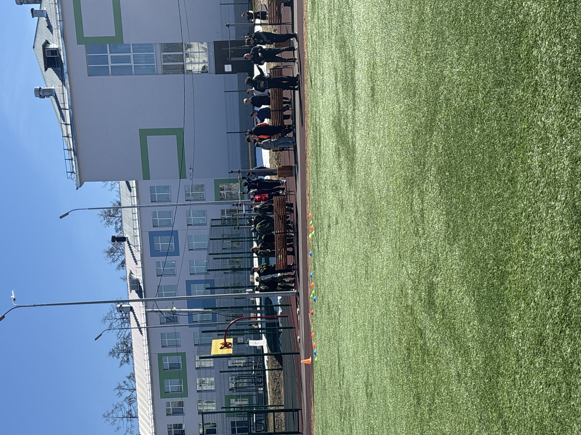 The image shows a group of people, some in military uniforms, standing and sitting on benches next to a green artificial turf field. The people appear to be observing something off-camera to the right. Orange and blue cones are arranged along the edge of the field. A basketball hoop and fenced area are visible. Buildings with green and blue accents are in the background. Based on the location mentioned in the prompt, this scene is likely located in the city of Chaikovsky, Russia.
