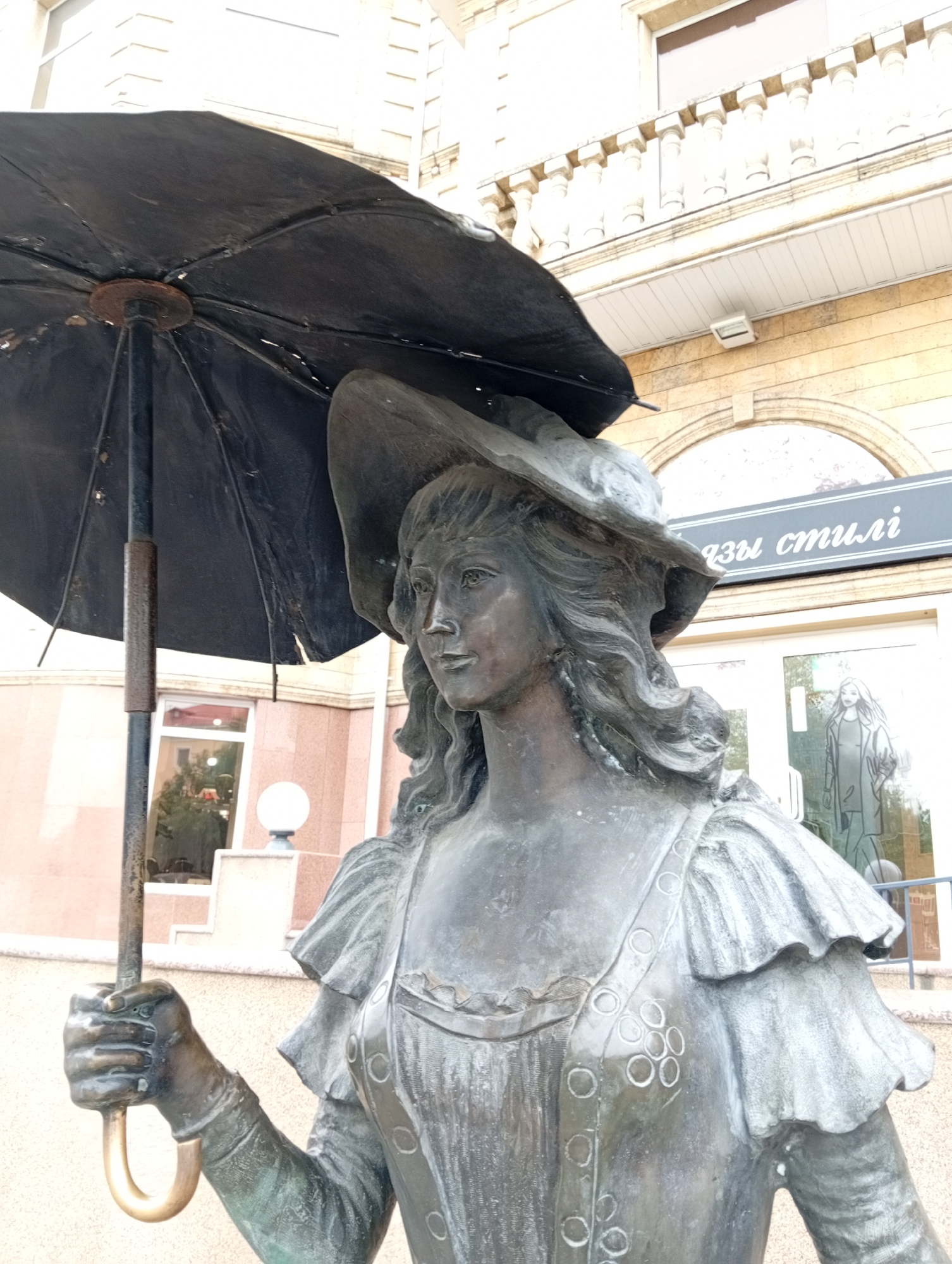 A weathered bronze statue of a woman holding an umbrella stands in front of a building with light-colored stone facade. The woman wears a wide-brimmed hat and a dress with ruffled sleeves. The building's facade features classical architectural elements, including columns and a decorative frieze. Above a glass storefront, a sign with Cyrillic lettering is visible. The ground is paved, and a lamppost with a spherical light fixture is situated to the left of the statue. The statue is located in the city of Kostanay, Kazakhstan.