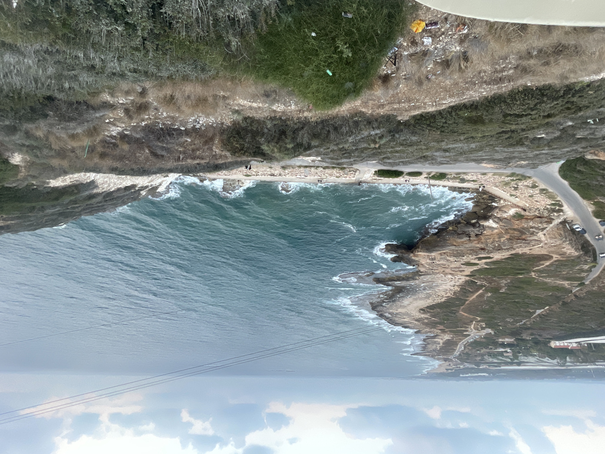 The image captures a scenic view of the Mediterranean coastline in the Mate Asher Regional Council, Israel. A winding road snakes along the rocky shoreline, with cars visible on it. The sea is a vibrant turquoise, with waves crashing against the rugged cliffs. Vegetation, a mix of dry grasses and green shrubs, covers the hillside in the foreground, with some scattered litter visible on the ground. In the distance, a city skyline is faintly visible on the horizon. The sky is overcast with scattered clouds, suggesting a possibly cool or breezy day. A few people are seen walking along the coastal path and on the beach area. The overall scene portrays a natural landscape being encroached upon by human development.