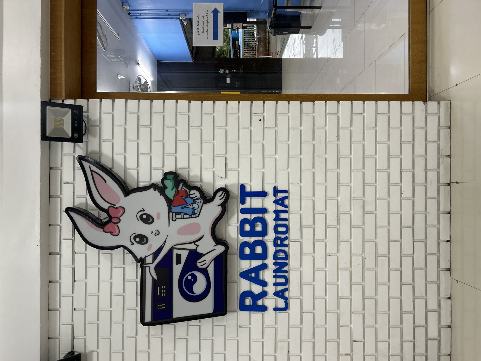 The image shows the exterior of a laundromat called "RABBIT LAUNDROMAT" located in Khlong Luang Town Municipality, Thailand. The laundromat features a white brick wall with a large, cartoonish decal of a white rabbit wearing a pink bow, holding a basket of clothes next to a washing machine. Below the decal, the name of the establishment is displayed in large blue letters. To the right of the laundromat's entrance, a blue directional sign with white text and an arrow pointing to the right is visible, indicating the way to the customer service area or reception. The entrance appears to be a doorway leading into the laundromat.