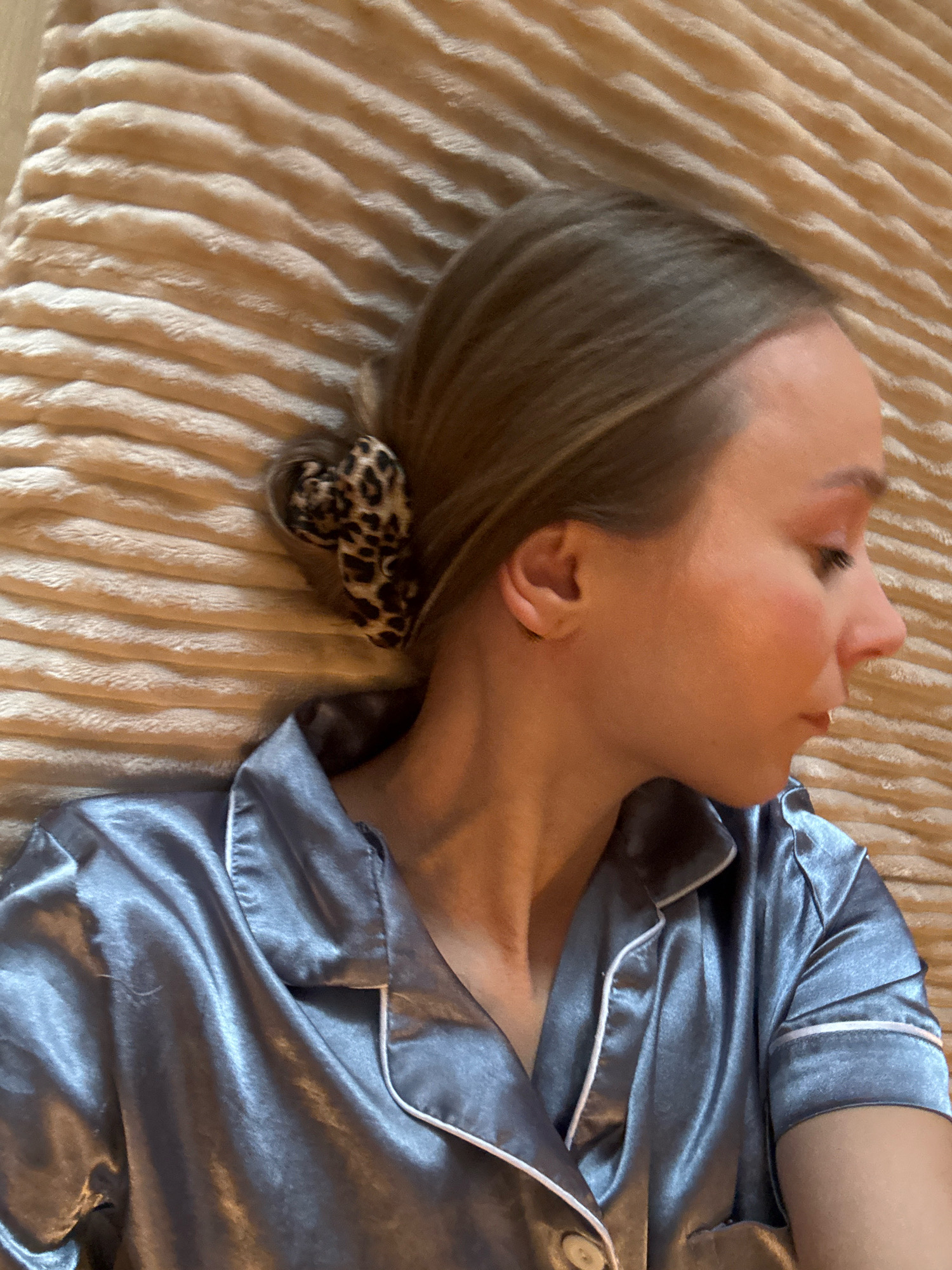 A close-up, top-down shot shows a woman lying down. Her head is turned to her right, profile visible, and her eyes are closed. She has blonde hair styled in a bun with a leopard print scrunchie. She is wearing a light blue satin pajama top with white piping. The background is a textured, beige blanket. The scene is indoors. There is no information to suggest this image is located in Überherrn, Germany.