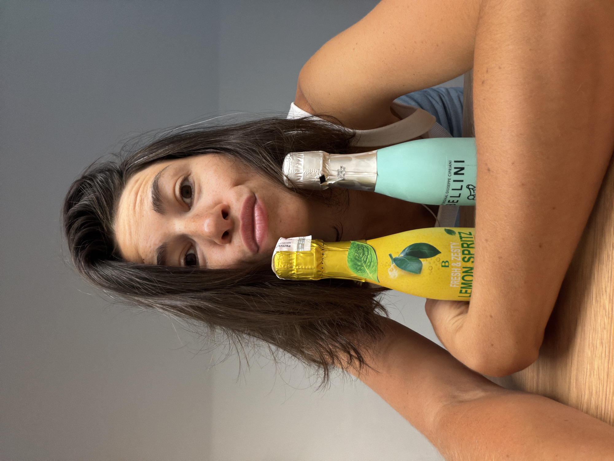 A woman with dark hair, puckered lips, and brown eyes is seated at a wooden table, holding two small bottles of sparkling wine. The bottle on the left is yellow and labeled "B FRESH & ZESTY LEMON SPRITZ" with an image of lemons. The bottle on the right is turquoise and labeled "BELLINI ORIGINAL GIUSEPPE CIPRIANI." Her arms are resting on the table, framing the bottles. The background is a plain grey wall. The scene appears to be indoors, possibly in Budva, Montenegro.