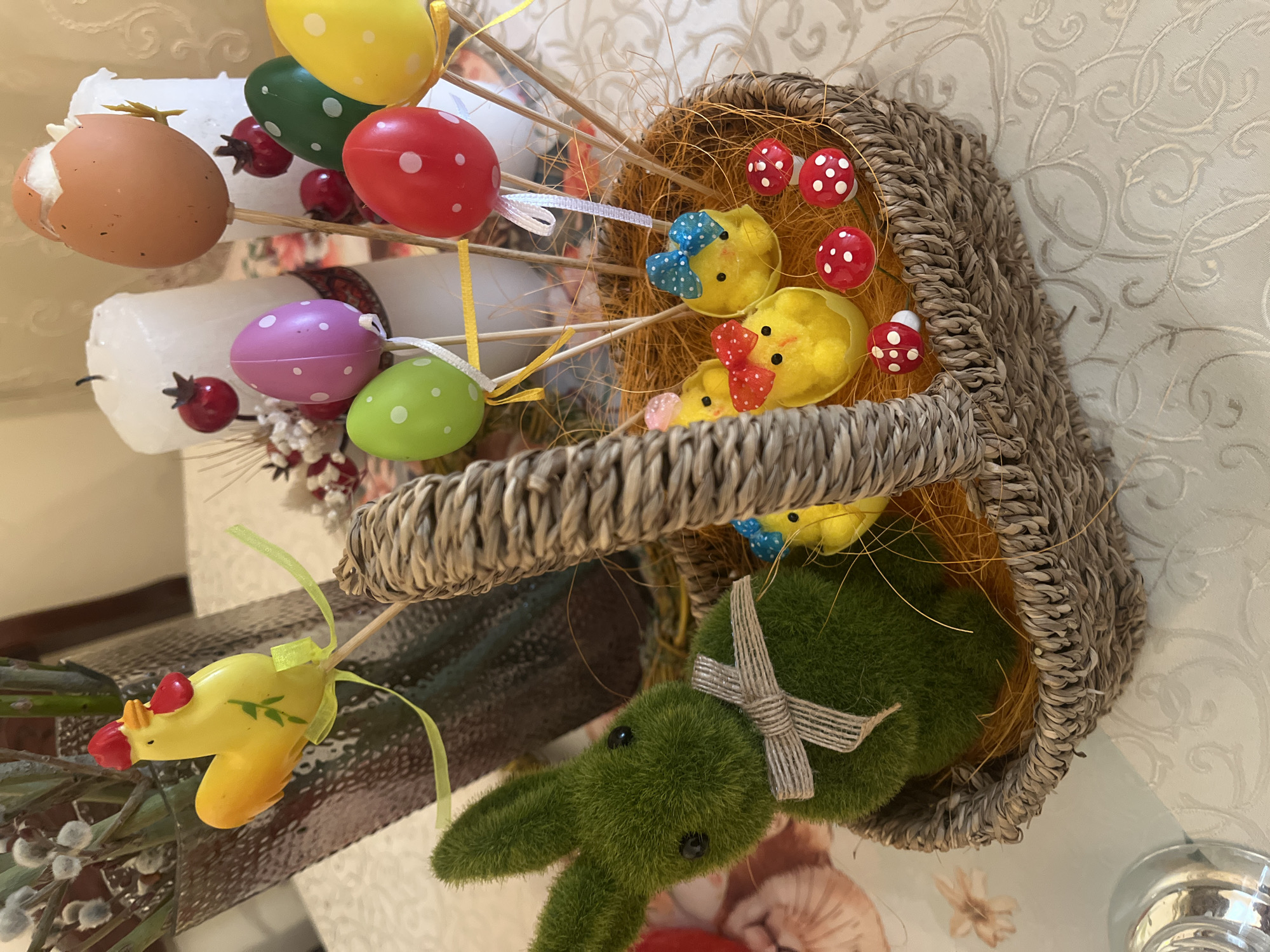 The image shows an Easter decoration. The foreground contains a wicker basket filled with artificial straw, small yellow chicks with bows, and red and white polka-dot mushrooms. A green, moss-covered bunny with a ribbon is positioned near the edge of the basket. In the background, there are white candles with decorative eggs on sticks in various colors (yellow, red, green, purple, brown). The scene is set on a patterned surface, possibly a tablecloth. The location is not evident from the image.