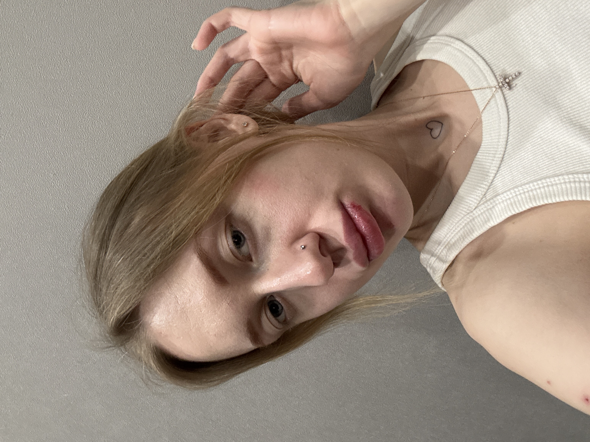 The image displays a close-up, chest-up view of a light-skinned female subject, captured indoors against a textured light-grey wall. The subject has blonde hair, parted to her left, and is looking directly at the camera with her head slightly tilted to her right. Her right hand is raised, with fingers near her right ear, appearing to touch or adjust her hair.
She wears a white ribbed tank top. A delicate gold-colored chain with a diamond-studded cross pendant is visible around her neck. A small, black outlined heart tattoo is located on the front of her neck, slightly to the left of the center. A silver-colored stud piercing is present on the left side of her nose, and a small, light-colored stud earring is visible in her right earlobe. Her lips appear full, with a noticeable reddish lesion or dried blood present on her lower lip. Two small, reddish marks are evident on her left upper arm/shoulder area.
The specific location within Rostov-on-Don, Russia, cannot be determined from the visual information provided in the image.