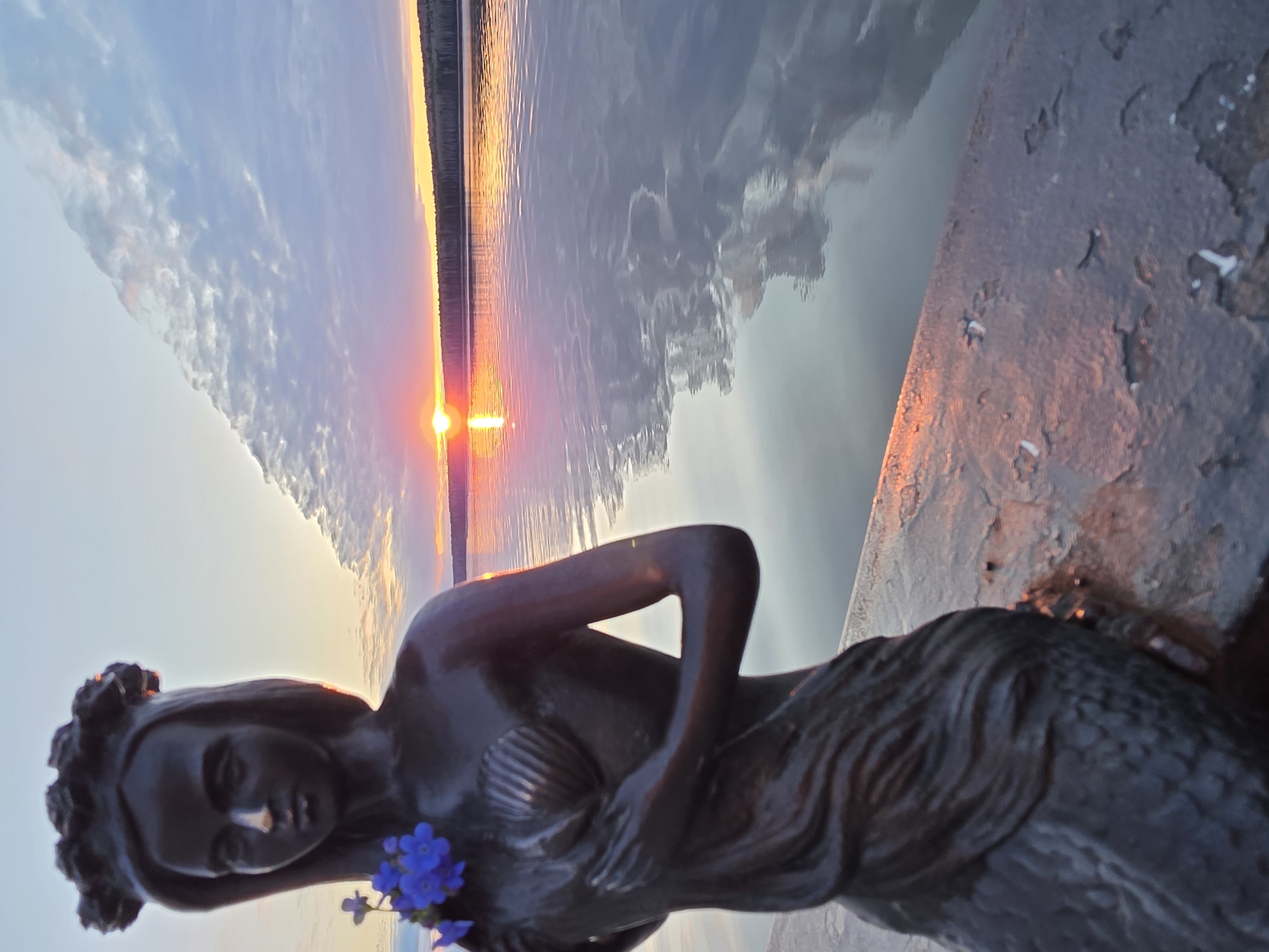 Here is a description of the image:
The image shows a bronze mermaid statue adorned with blue flowers, lying on a concrete embankment with a body of water in the background. A sunset with orange and pink hues is visible on the horizon, reflecting off the water's surface. The sky above the horizon is a mix of blue and cloud cover. The image is taken at an angle, making the horizon appear diagonal. The location is the city of Chaikovsky, Russia.