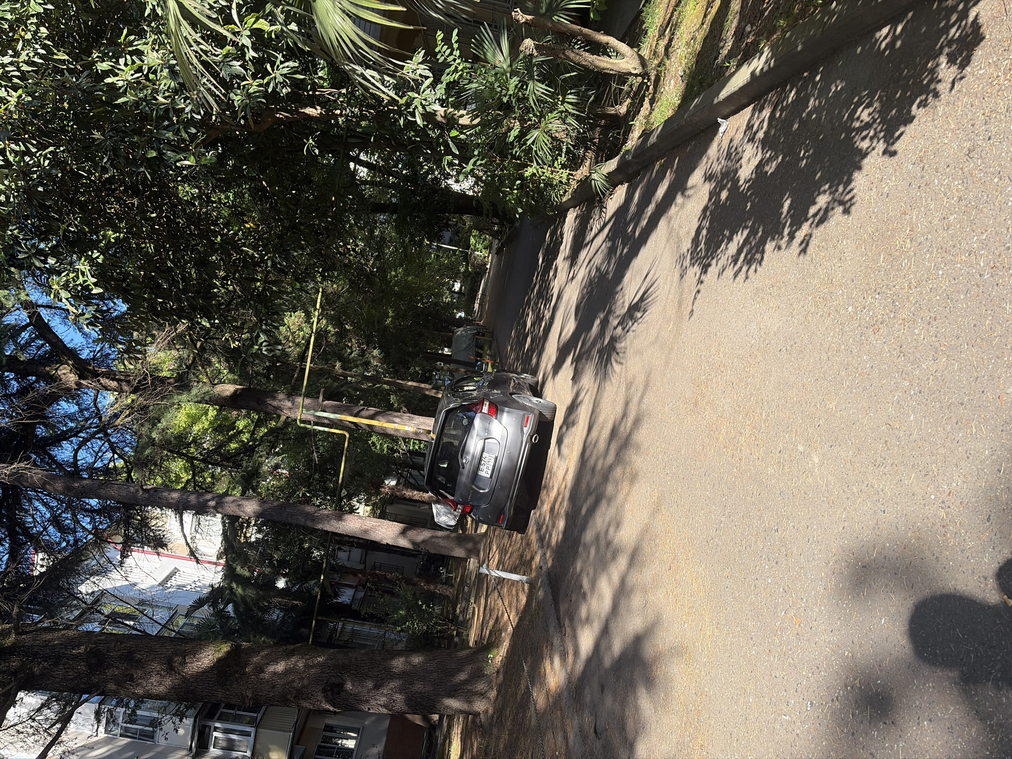 The image depicts a street scene in Sochi, Russia, characterized by a residential building and abundant vegetation. A grey hatchback car is parked on the right side of the asphalt road, facing away from the viewer. The car's license plate is partially visible, reading "E 574 PP 193". Trees with dense foliage line both sides of the street, casting shadows on the ground. A white fence post is visible on the left side of the road, near the parked car. The building on the left appears to be an apartment complex with multiple windows and balconies. The sky is blue and visible through the tree branches. The overall impression is that of a sunlit, residential area within a city that has significant green spaces.