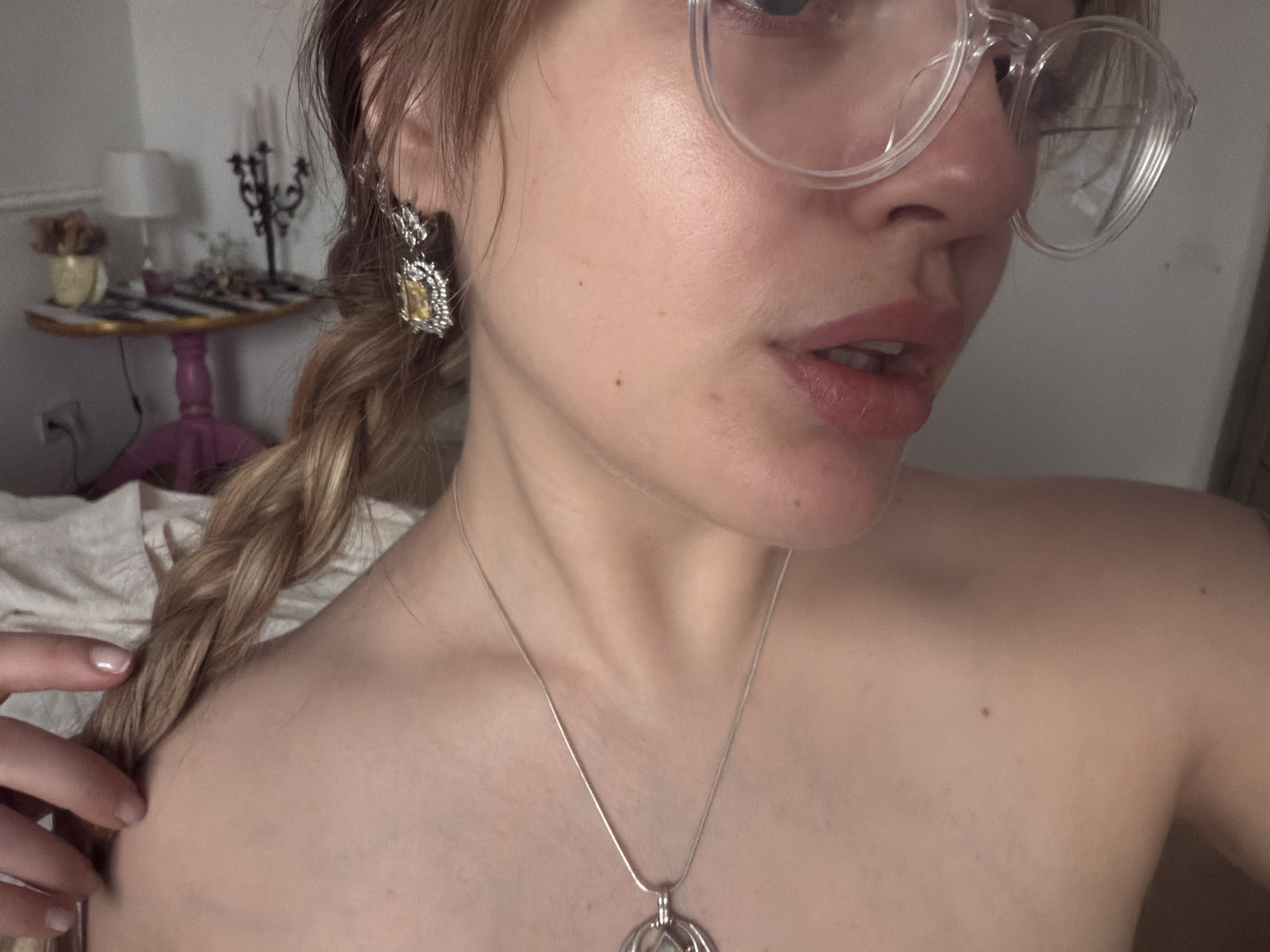 A close-up shot shows a fair-skinned person with a blonde braid, wearing transparent-framed glasses and jewelry. The person is wearing an earring with a yellow stone and a silver necklace with a pendant. The background features a table with a lamp and decorative objects. The location is Больша́я Шильна́, Russia, based on available data. No actions or interactions are visible.