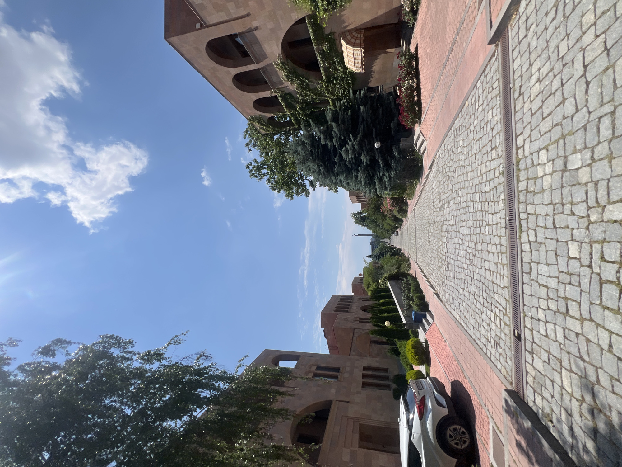 The image captures a cobblestone pathway lined with identical stone buildings and lush greenery, extending towards a distant monument against a clear blue sky. The architecture suggests a well-maintained, possibly historical or institutional, setting within the city of Yerevan, Armenia. The sun shines brightly, casting shadows and highlighting the textures of the stone and paving. On the left, a white SUV is parked near one of the buildings. The pathway is slightly sloped, leading the eye towards the monument, which appears to be a tall, slender structure. The buildings on either side feature arched windows and balconies, some adorned with climbing ivy. The scene conveys a sense of order and tranquility, with no people visible in the frame. The overall impression is that of a serene and stately urban landscape.
