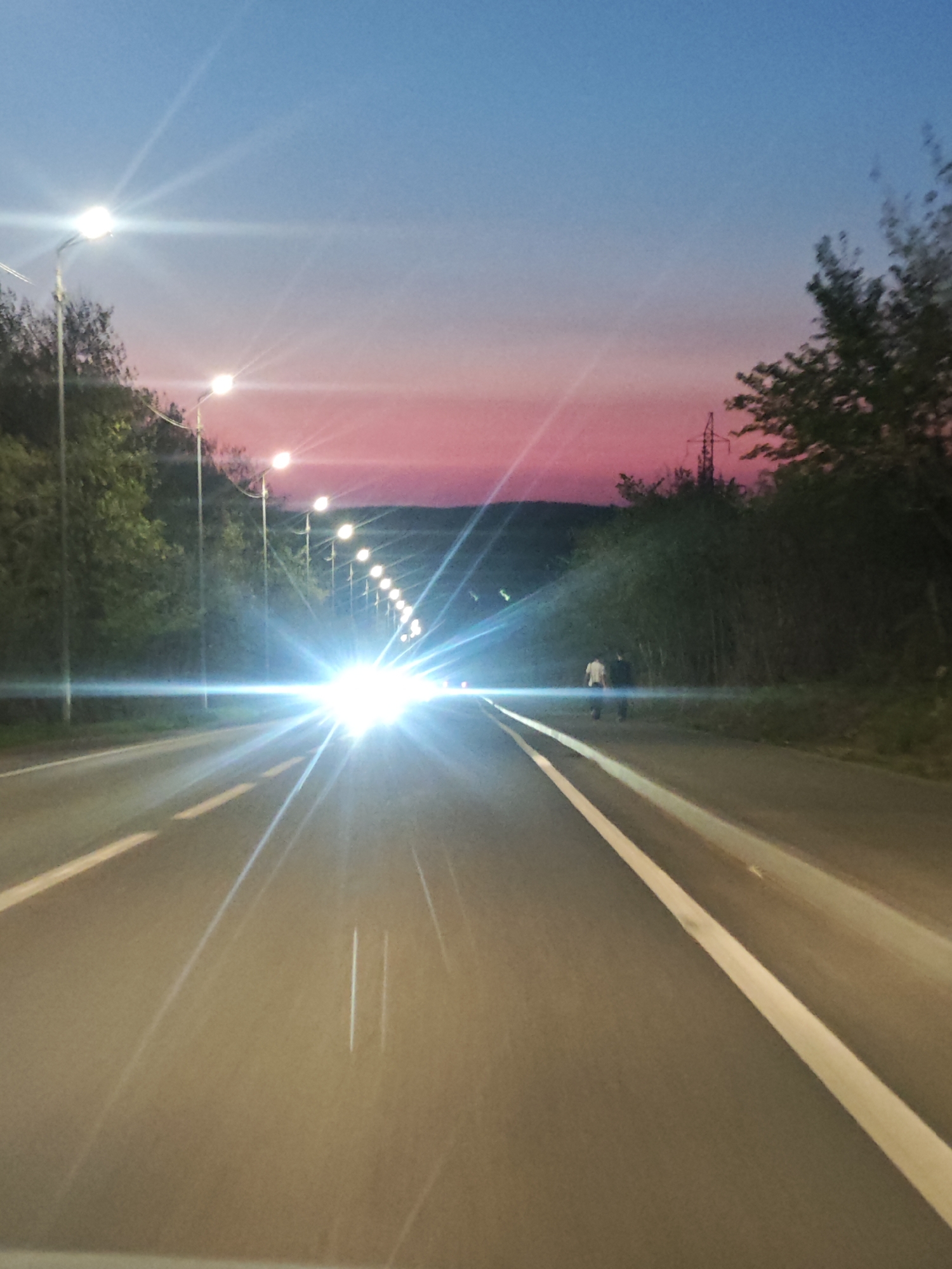 The image depicts a road at dusk, possibly near Samara, Russia, indicated by the power lines and streetlights. The sky displays a gradient of blue to pink. An oncoming vehicle with bright headlights dominates the view. Two individuals are walking on the right shoulder of the road. Streetlights illuminate the road, which has white lane markings. Trees line both sides of the road.