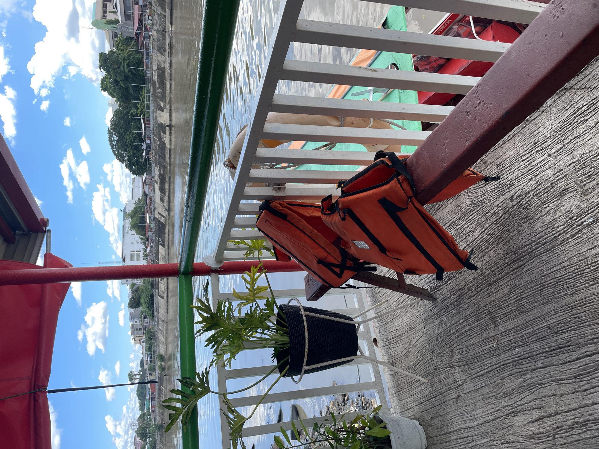 On a deck of a boat, two orange life vests are hanging on a red railing. The boat is on a river with a city skyline in the background. The sky is blue with scattered white clouds. The deck is made of concrete and has a potted plant.