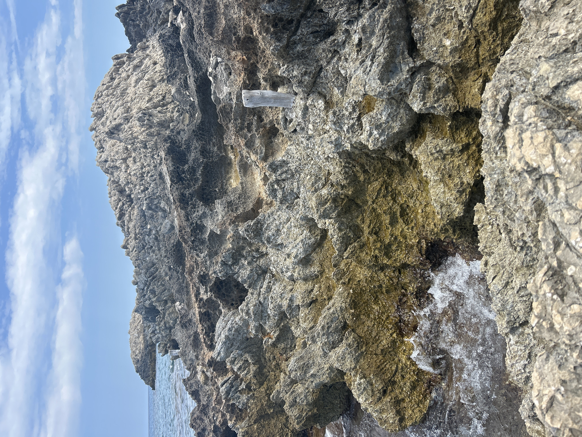 The image depicts a rugged, rocky coastline with the sea in the background. The foreground is dominated by rough, grey and brownish rocks, some of which are partially submerged in the water. Small waves are seen crashing against the rocks on the left side of the image. A weathered wooden post stands upright amongst the rocks in the middle ground. In the distance, the sea stretches towards a rocky outcrop, and a hazy blue sky with scattered white clouds completes the scene. The location is Cala Lliteres, Spain.