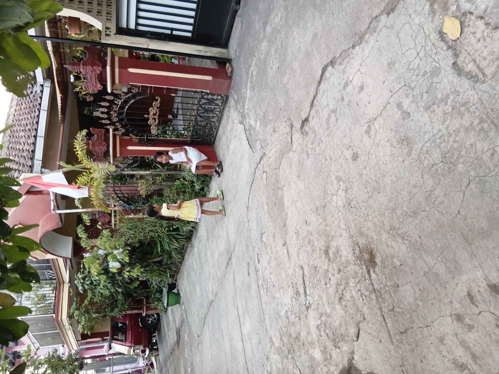 Two young girls are present in this outdoor scene. One girl, wearing a yellow dress and green sandals, stands on a concrete surface in front of a house. The other girl, dressed in a white dress and black shoes, sits on a concrete ledge next to a decorative metal gate. The house appears to be a residence in Tanggul Wetan, Indonesia. The property features a red car parked on the left side, surrounded by dense greenery. The foreground is a cracked concrete road, and the background consists of the house's facade, a gate, and a portion of a black metal gate. A red and white flag is visible on the house.