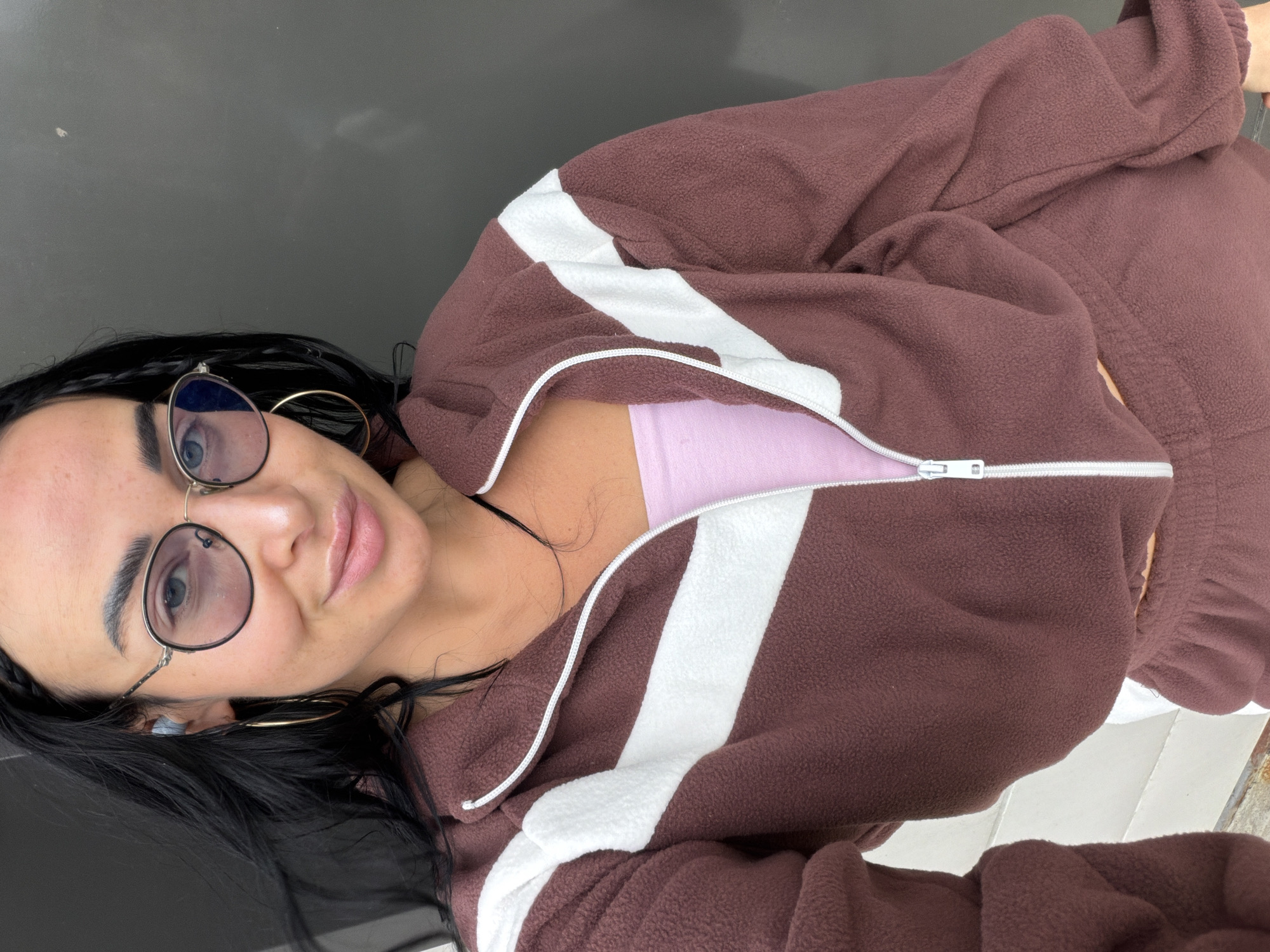 A young woman is posing for a close-up selfie outdoors. She has black hair, fair skin, and is wearing round glasses and gold hoop earrings. She is wearing a brown, fleece zip-up jacket with white stripes over a light pink top. The background appears to be a dark-colored surface, likely a wall or building exterior. The location is not discernible as Sust'e-Konets, Russia from the available image.