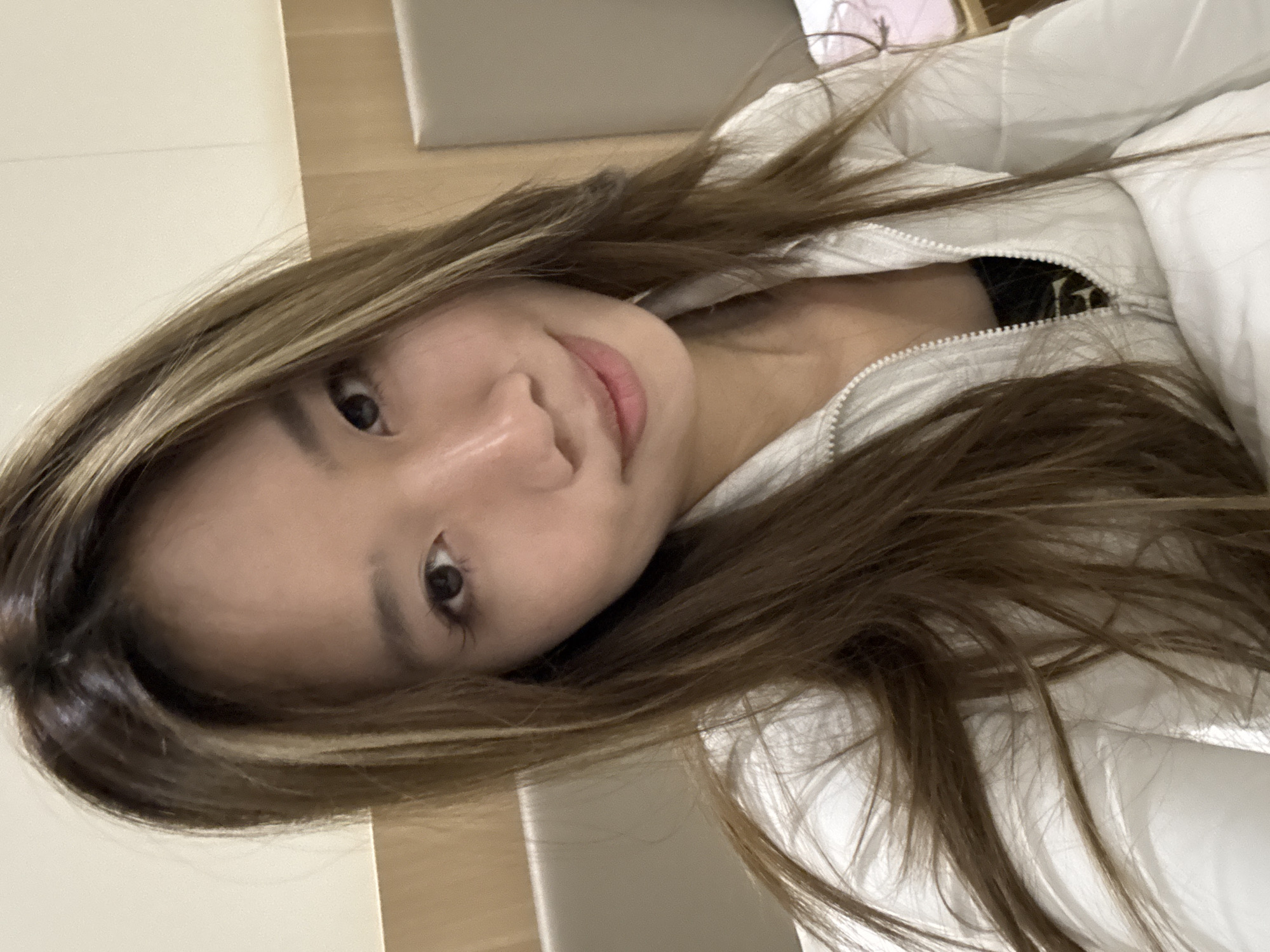 A close-up, slightly angled shot shows a woman with long, light brown hair with blonde highlights, wearing a white zip-up jacket. She appears to be indoors. No indicators of Dunhuang City, China, are visible in the image.