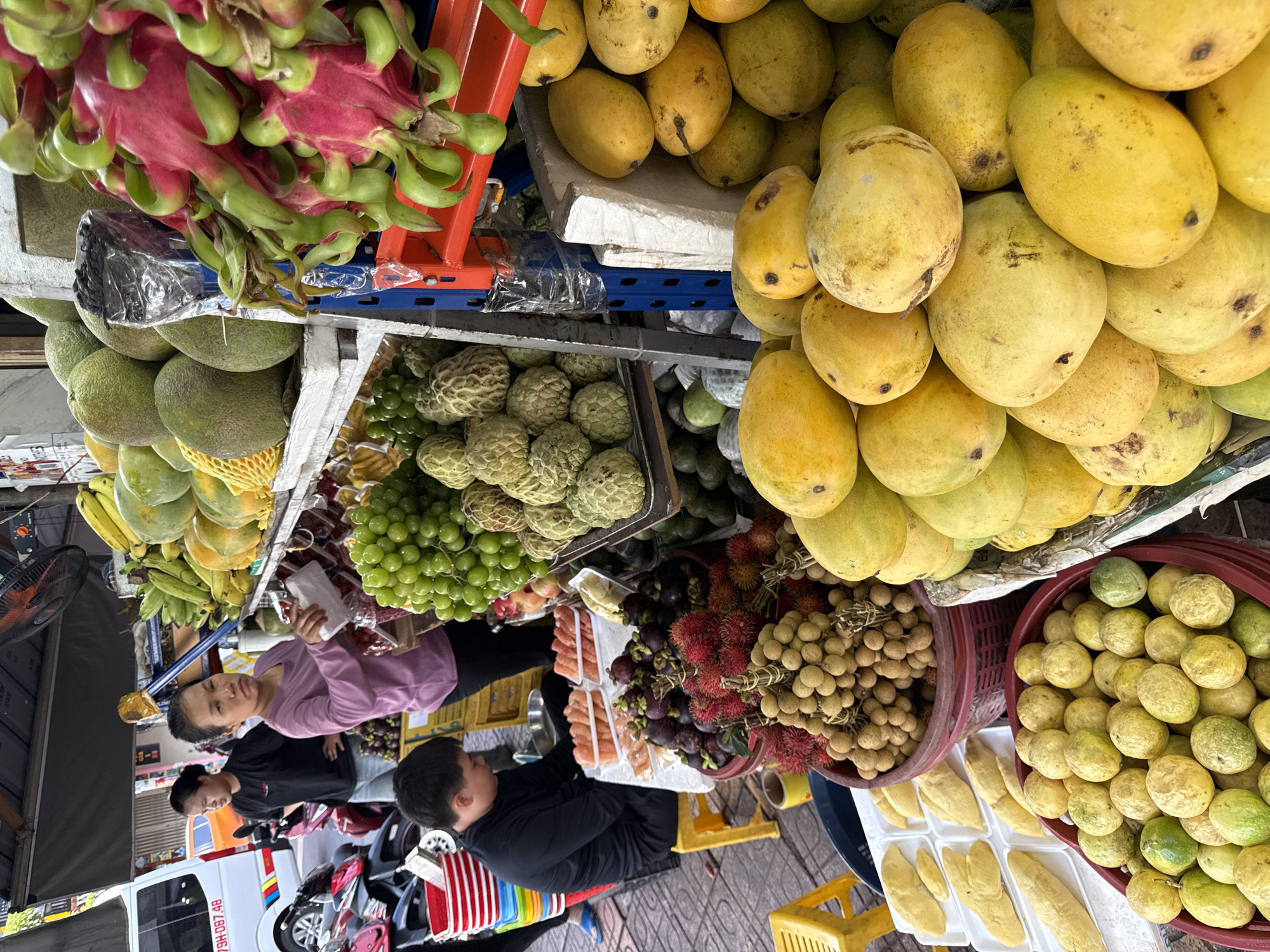 The image depicts a busy outdoor fruit stand in Nha Trang, Vietnam, likely a street market. A young person in a black t-shirt sits on a yellow stool, attending to a table with various fruits arranged in plastic containers. In the background, a woman in a purple shirt appears to be interacting with the stall, holding a white item. The stall itself is laden with a diverse selection of tropical fruits, including bunches of green grapes, dragon fruit, custard apples, mangoes, rambutan, longan, and passion fruit. Several motorcycles are parked nearby, and a white van is visible in the far background, suggesting an urban street setting. The ground is paved with cobblestones.